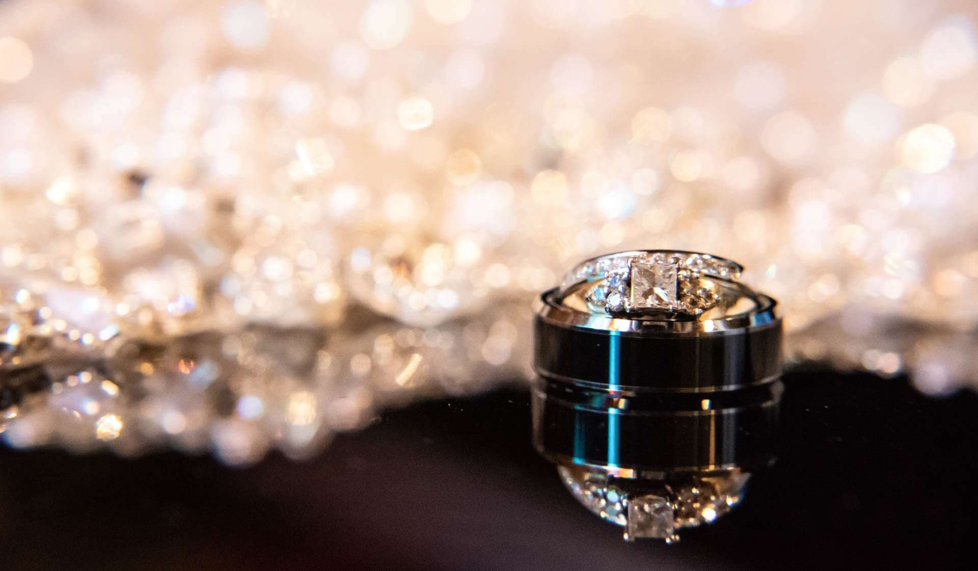 Diamond engagement ring stacked on a dark wedding band, reflected on glossy black with sparkling beadwork behind