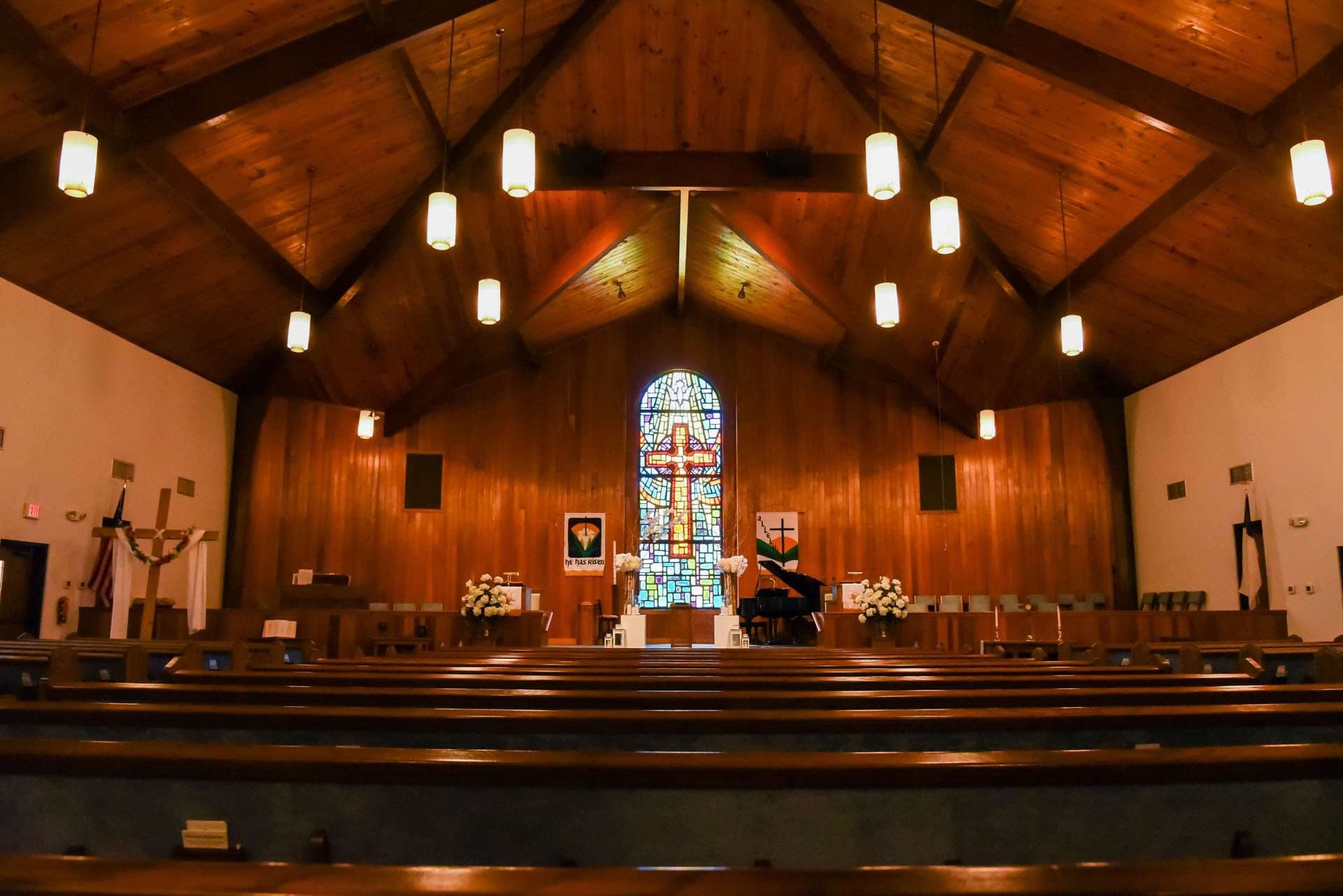 Empty church sanctuary with wood-paneled walls, rows of pews, and a stained glass cross window behind the altar