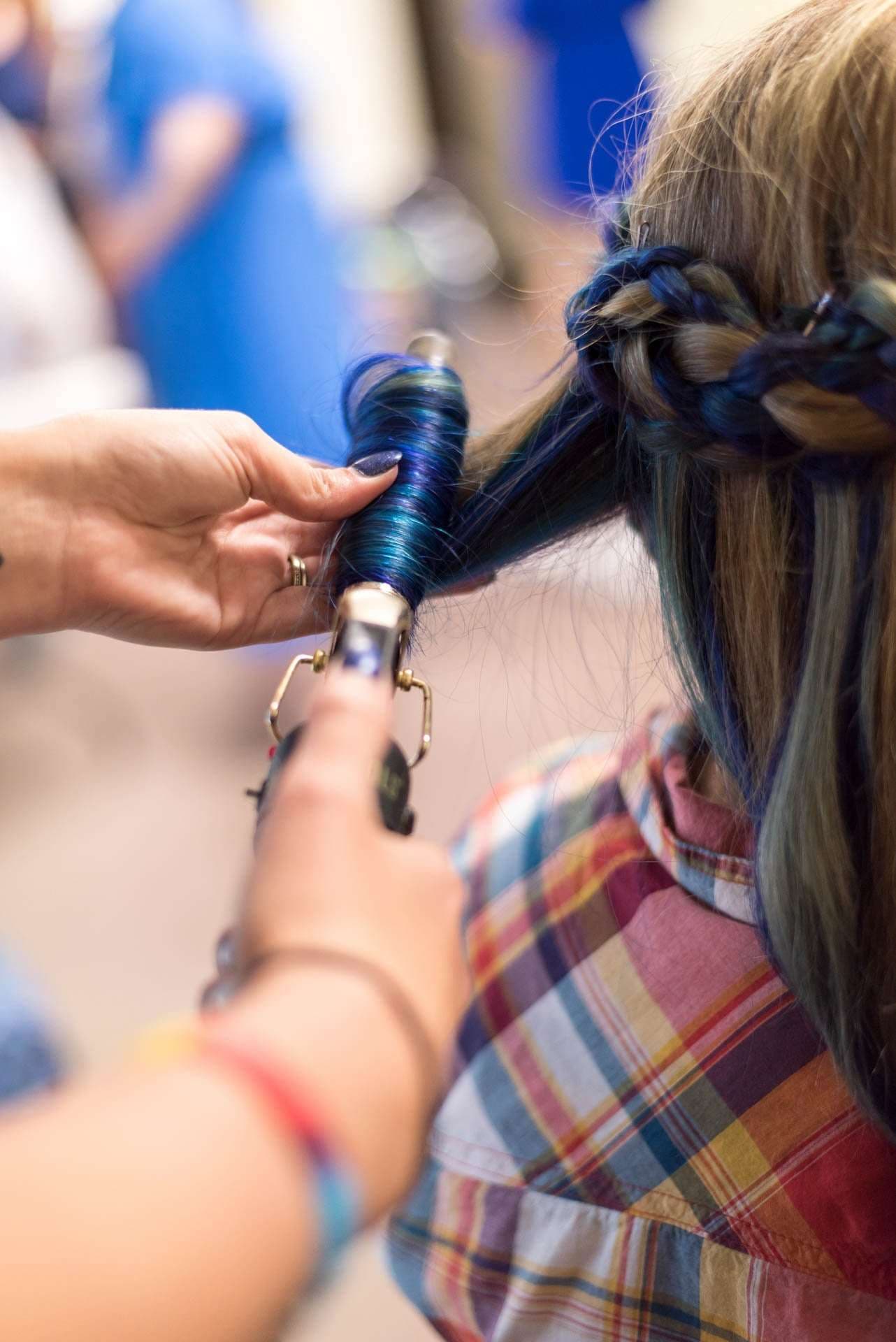 Hairstylist curling a section of blue-dyed hair with a curling iron beside a braided updo, client in a plaid shirt