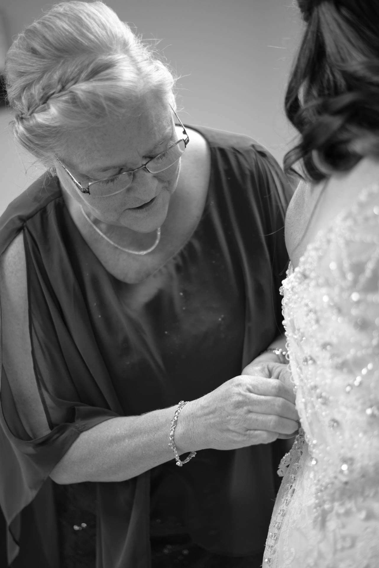Black-and-white image of an older woman with glasses fastening the beaded lace back of a bride's wedding gown