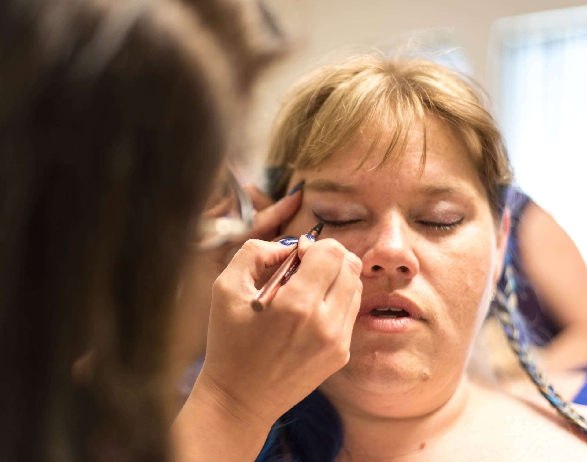 Makeup artist applying eyeliner to a woman with closed eyes and blonde bangs during wedding preparations