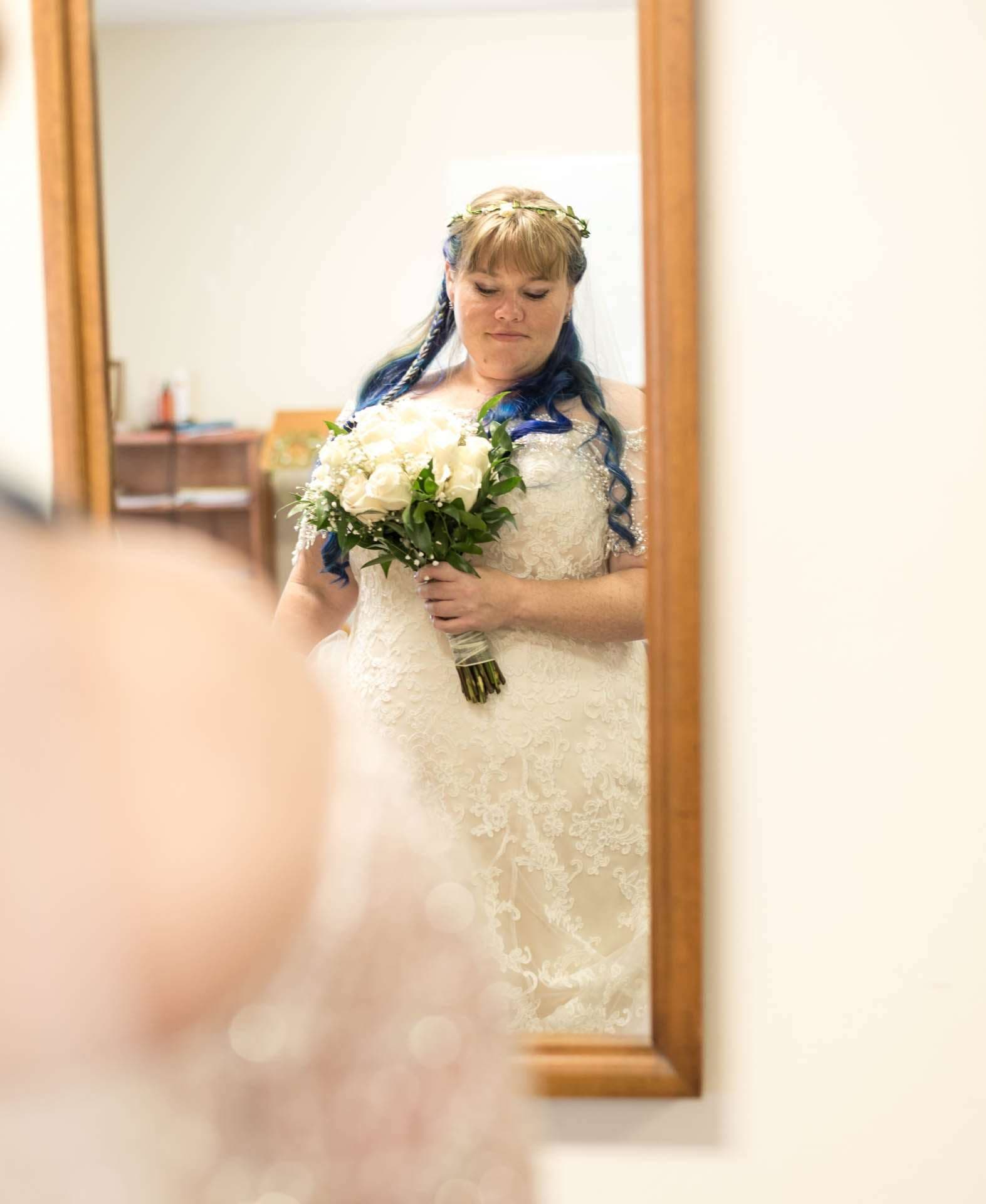 Bride with blue braided hair and a gold floral crown holds a white rose bouquet, reflected in a wooden-framed mirror