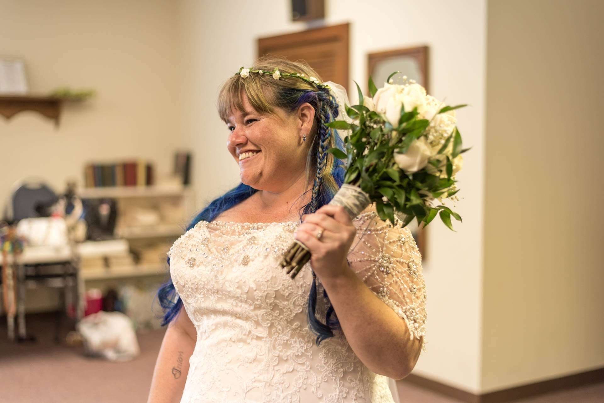 Smiling bride with blue-streaked braided hair and flower crown holds a white rose bouquet in a lace beaded gown
