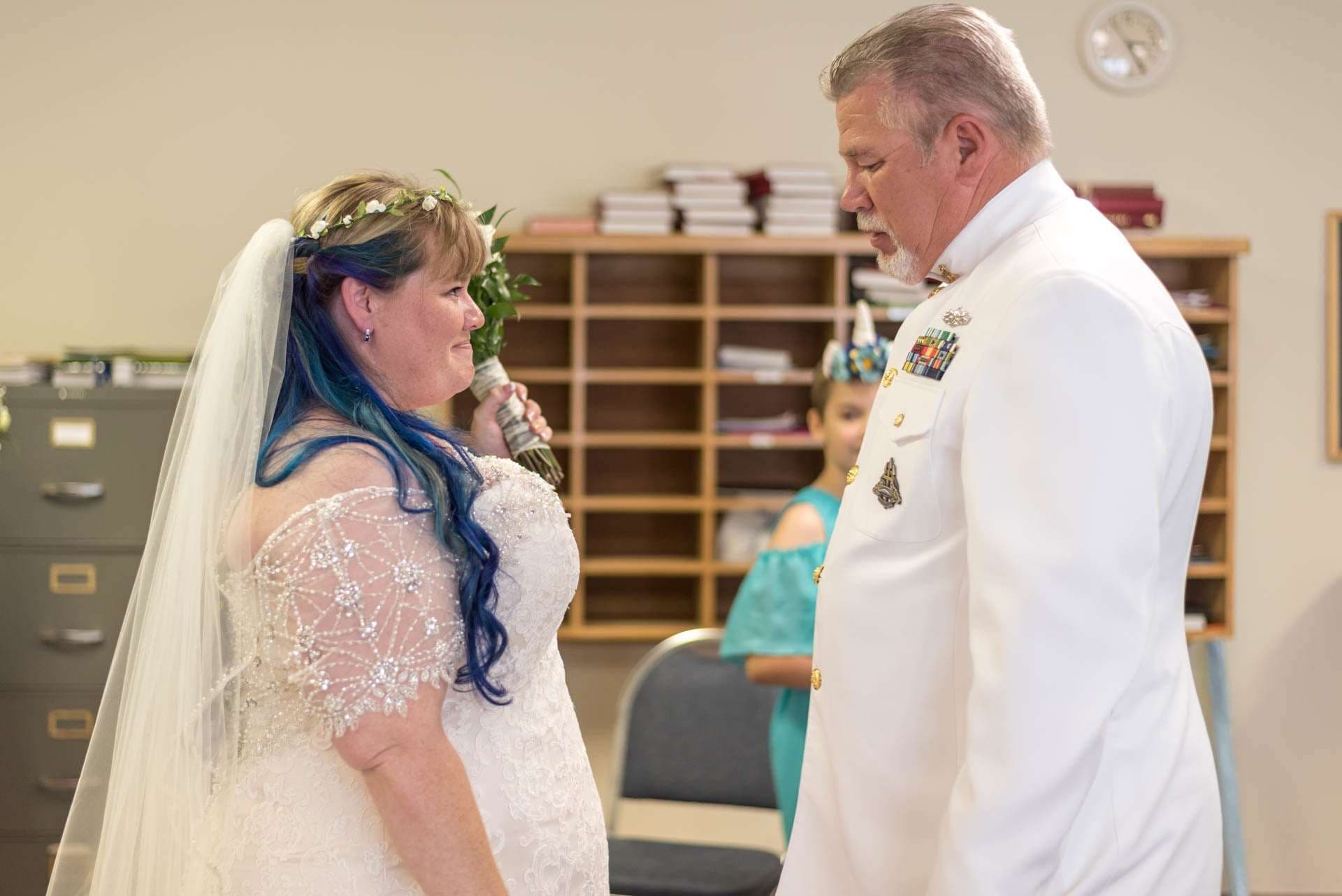 Bride with blue-streaked hair, flower crown and veil smiles at a groom in a white naval uniform inside a room