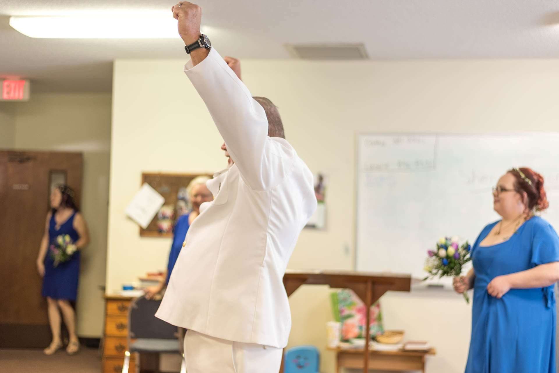 Man in white suit raising his fist in celebration while a woman in a blue dress holding a bouquet watches