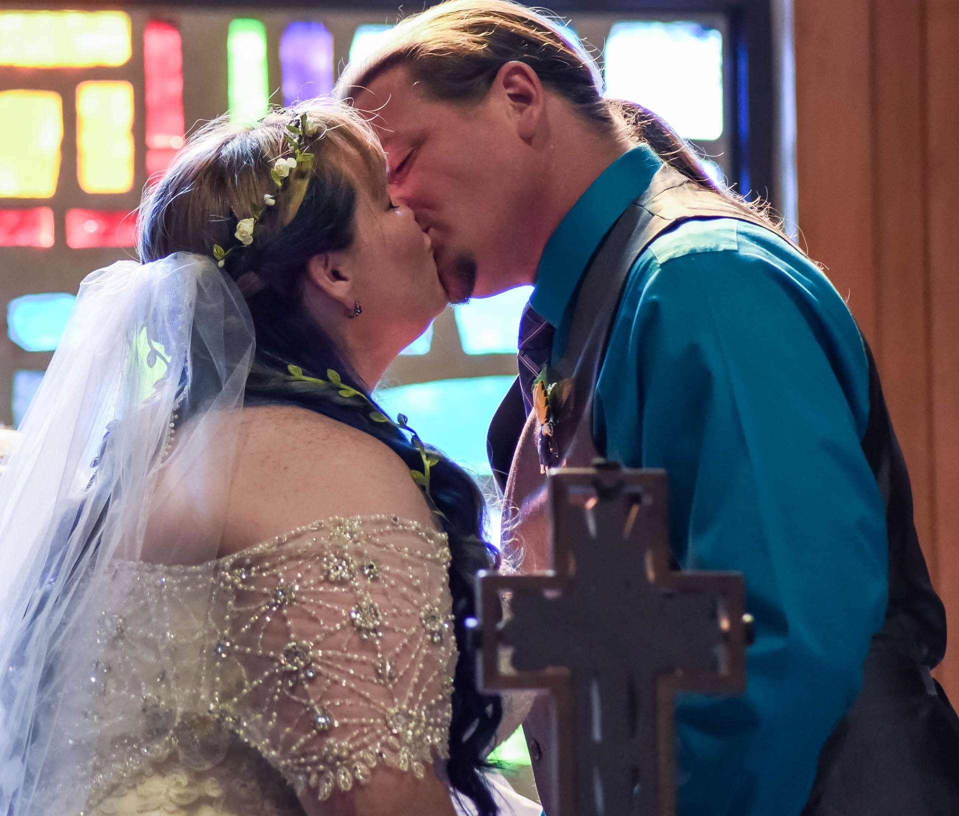 Couple kissing at a church altar beside a wooden cross, bride in a beaded gown and veil before stained glass