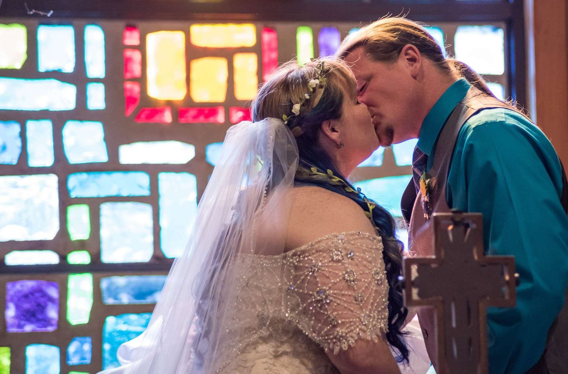 Couple kissing in front of a colorful stained glass window, the bride in a beaded lace gown and veil