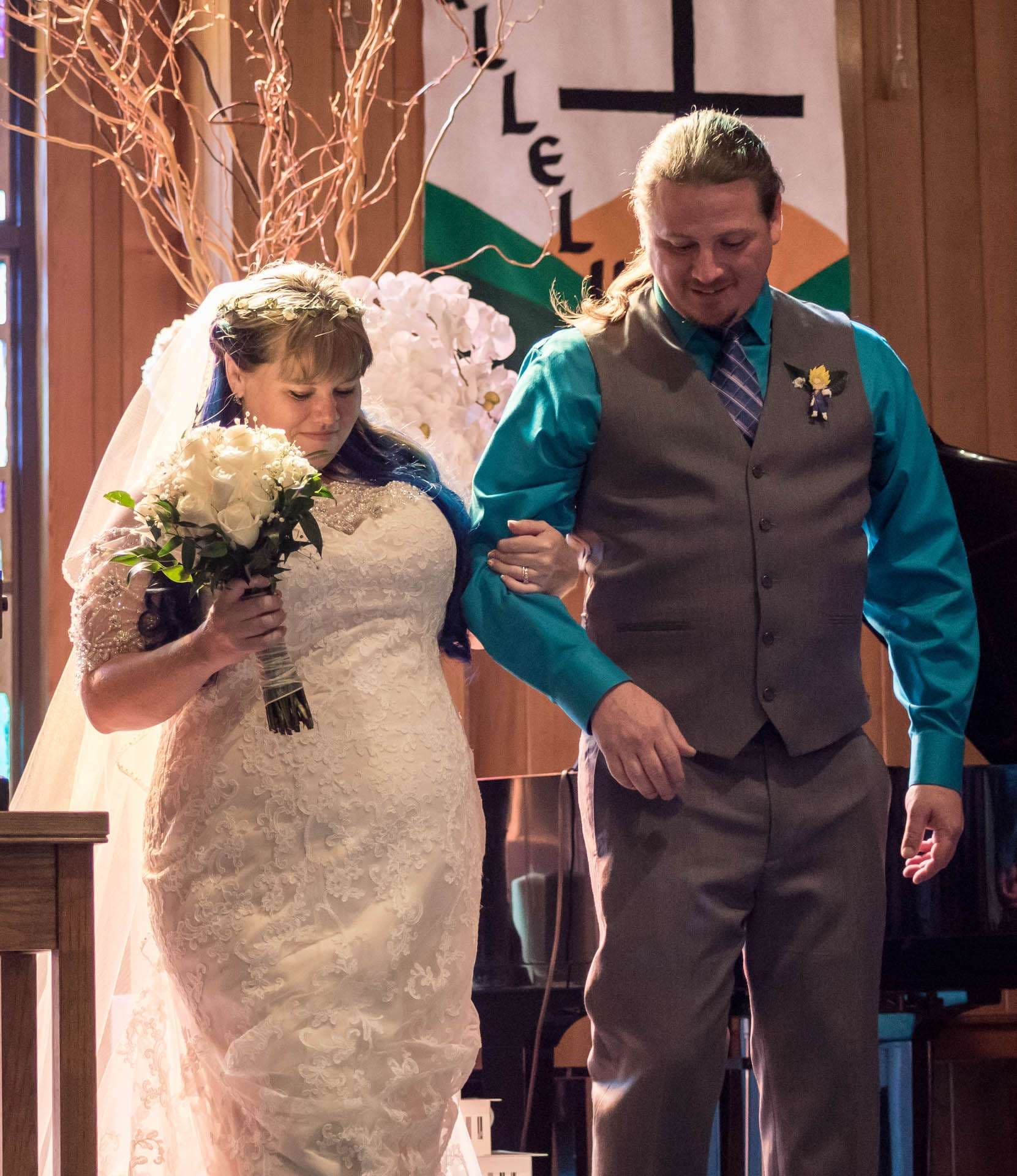 Bride in a white lace gown holding a white bouquet walks arm in arm with a smiling groom in a teal shirt and gray vest