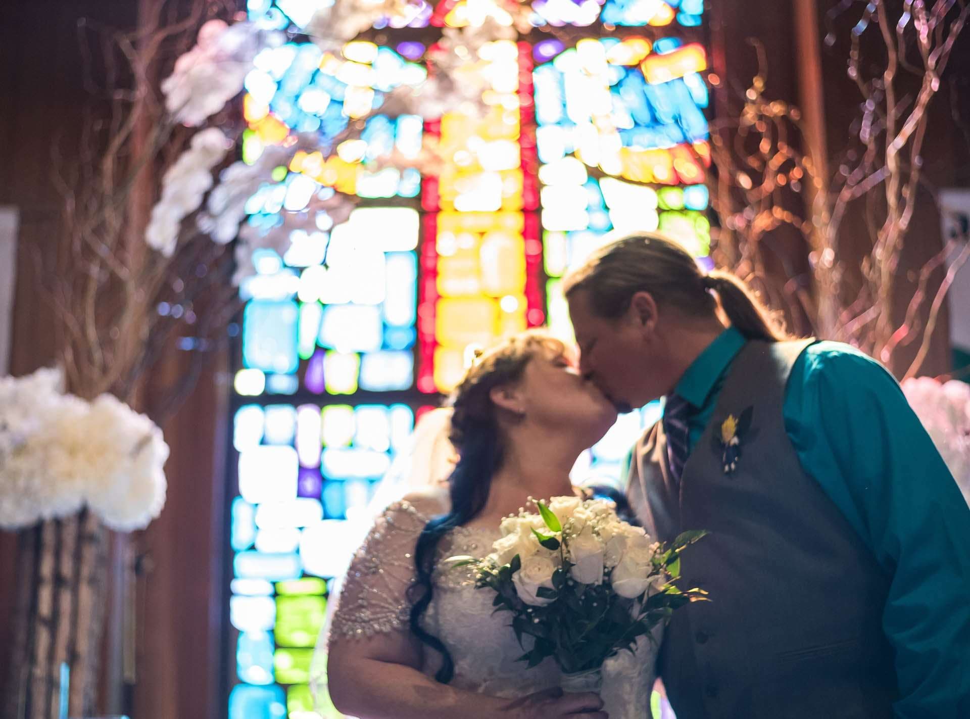 Couple kissing in front of a colorful stained glass window, the bride in a lace gown holding a white bouquet