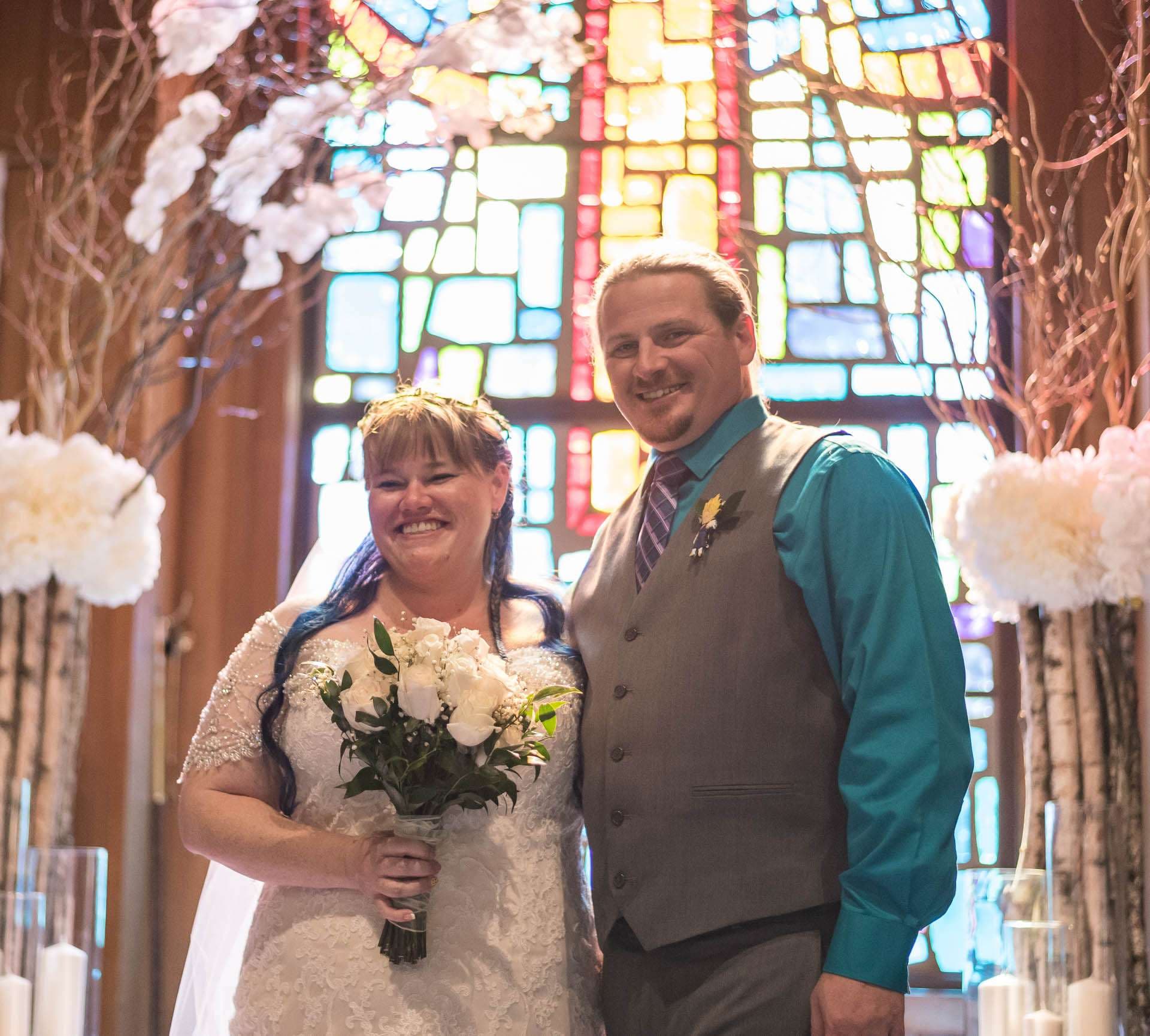 Smiling couple in front of a stained glass window, bride with blue-streaked hair holding white roses, groom in gray vest