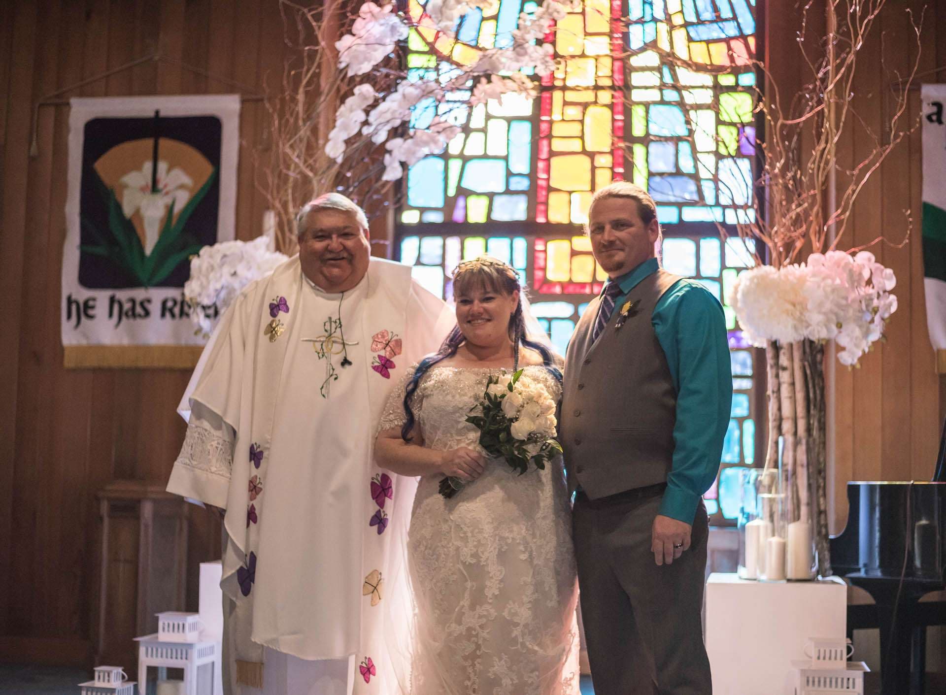 Smiling couple posing with an officiant in white vestments inside a church with stained glass and white floral arrangements