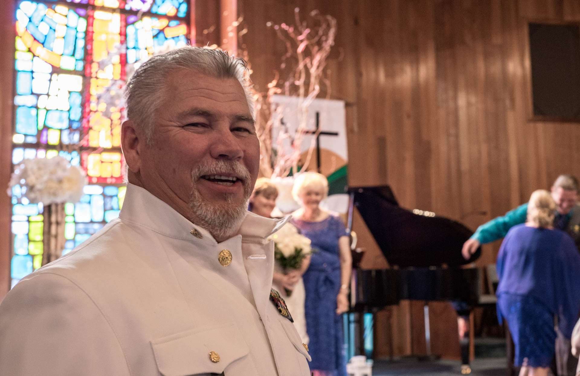 Smiling man in a white military-style dress uniform in a church with stained glass, guests and a piano behind him