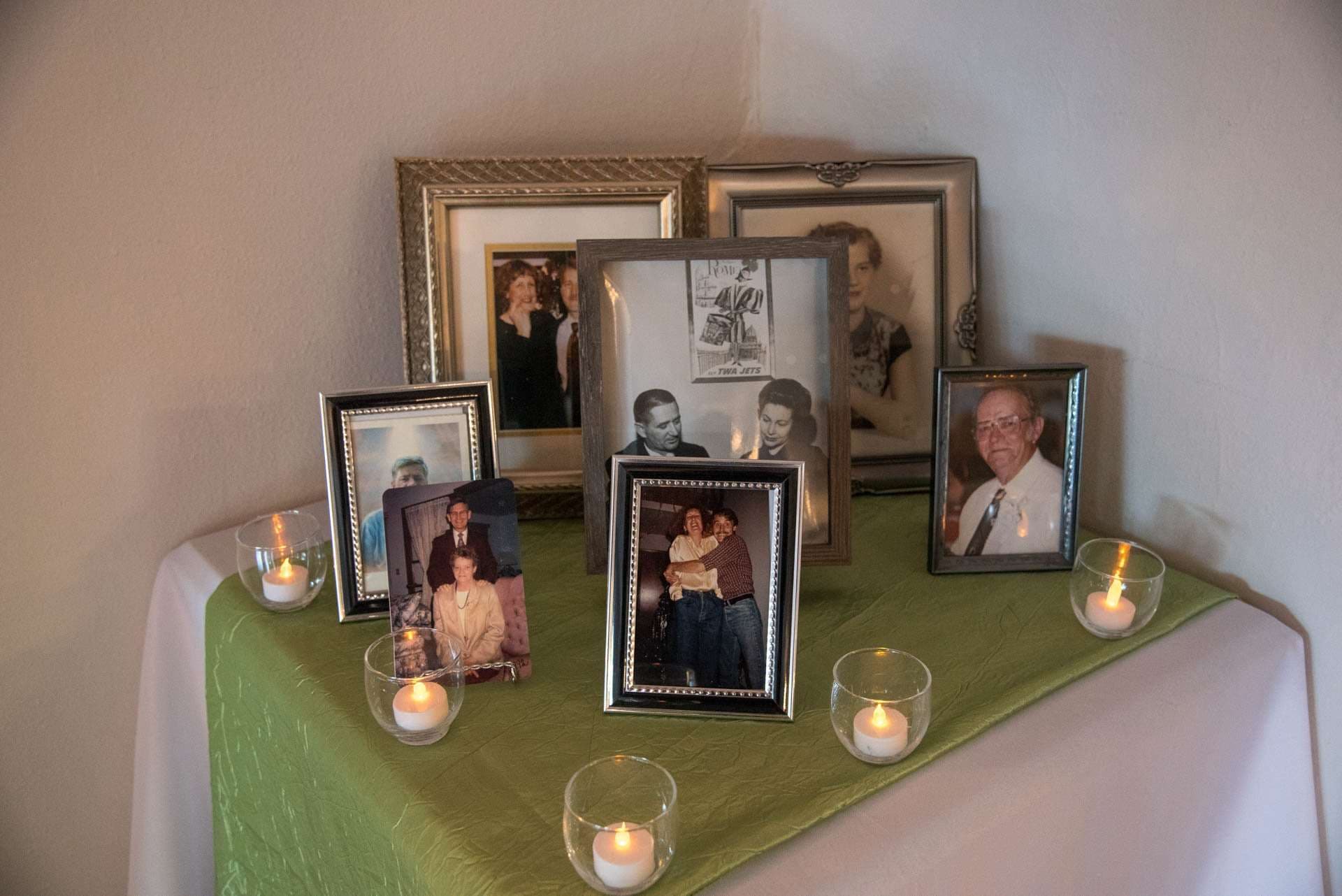 Framed family portraits, some black-and-white, displayed on a green-draped corner table with lit tea light candles