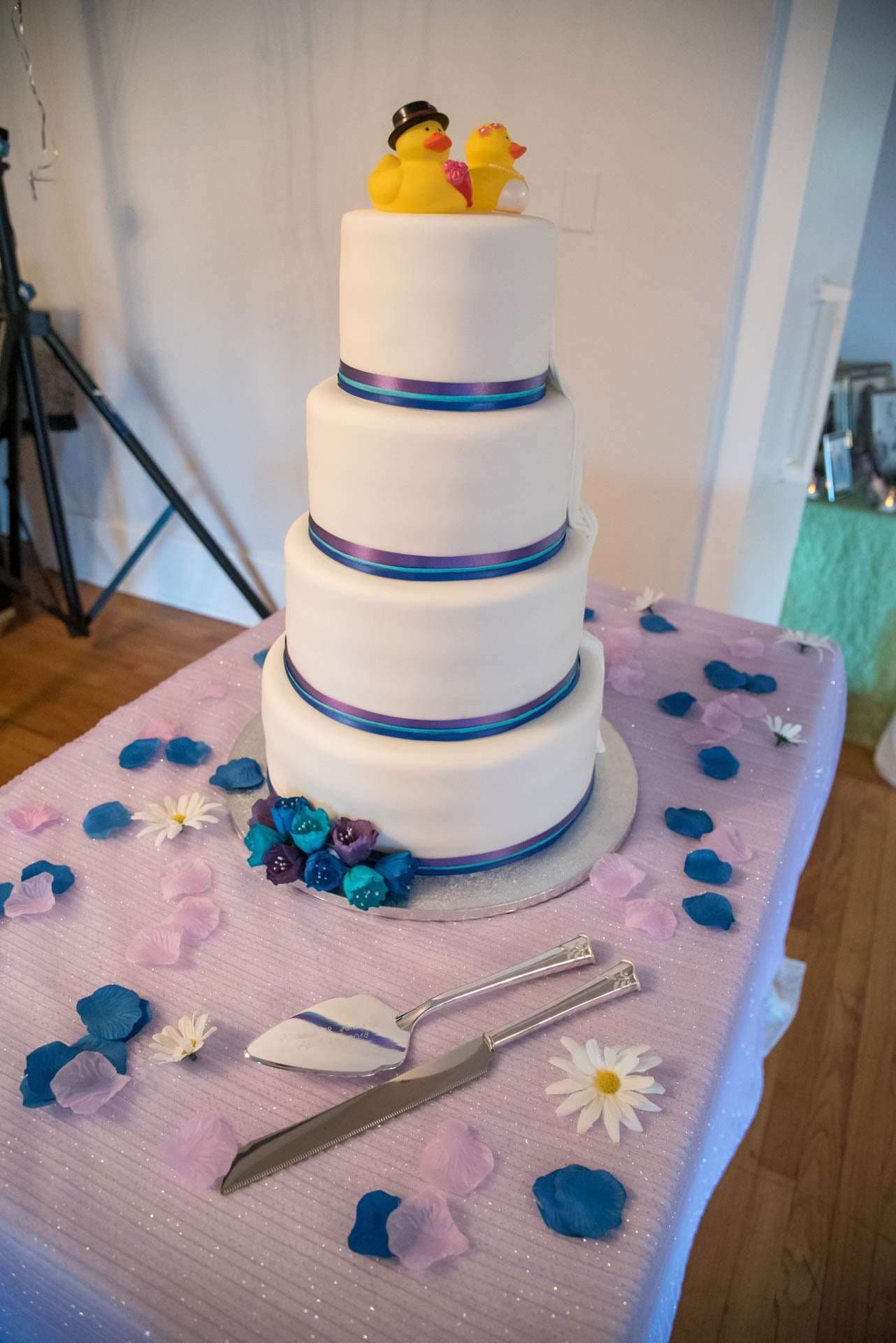 Four-tier white wedding cake topped with rubber ducks in a top hat and veil, beside a cake server and petals