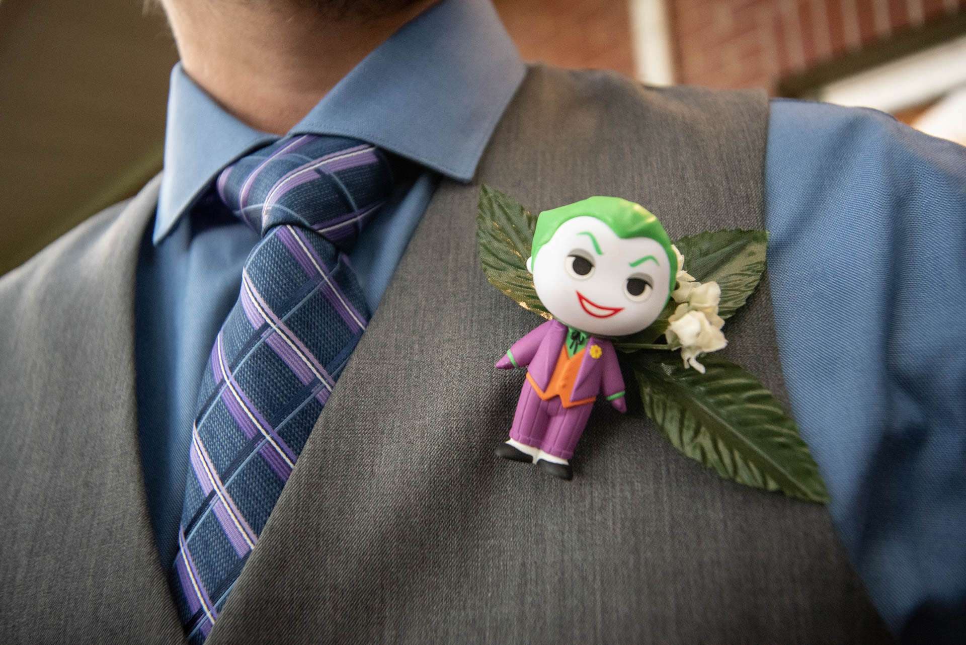 Joker figurine boutonniere with green leaves pinned to a gray vest, worn with a blue shirt and purple plaid tie