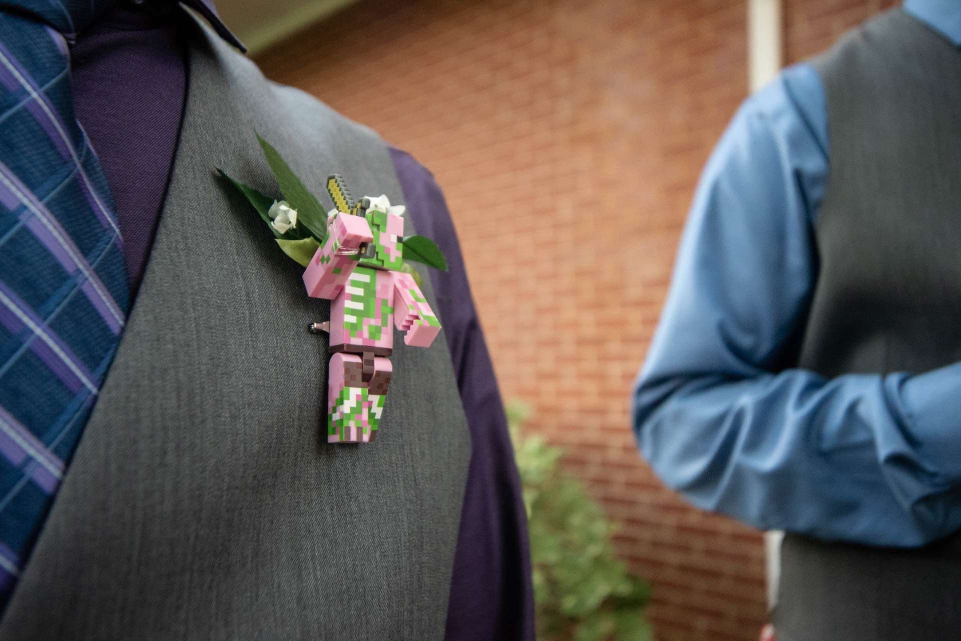 Pink pixelated Minecraft zombie pigman boutonniere with greenery pinned to a gray vest, another vested man blurred behind