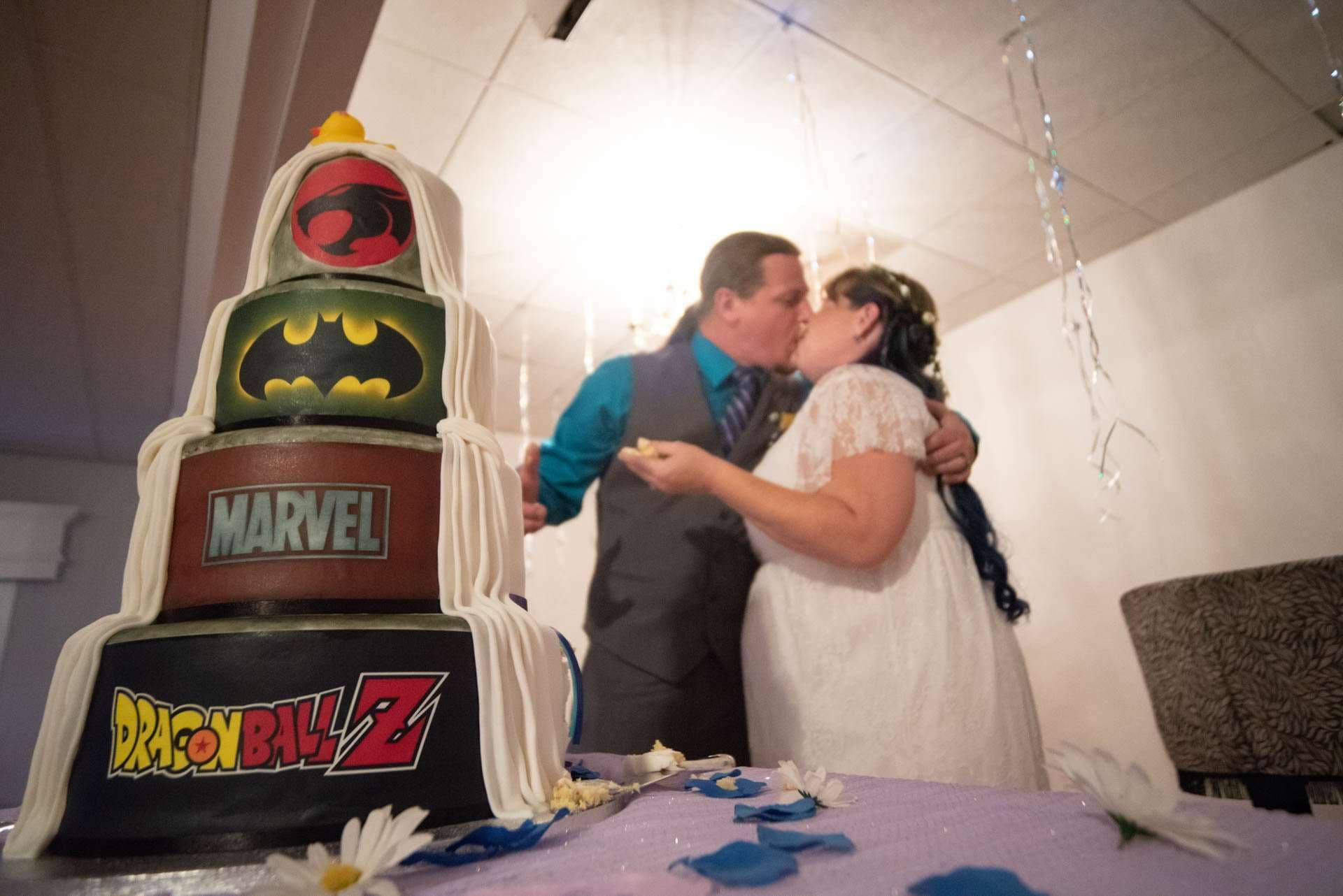 Couple kissing behind a tiered wedding cake decorated with Thundercats, Batman, Marvel and Dragon Ball Z logos