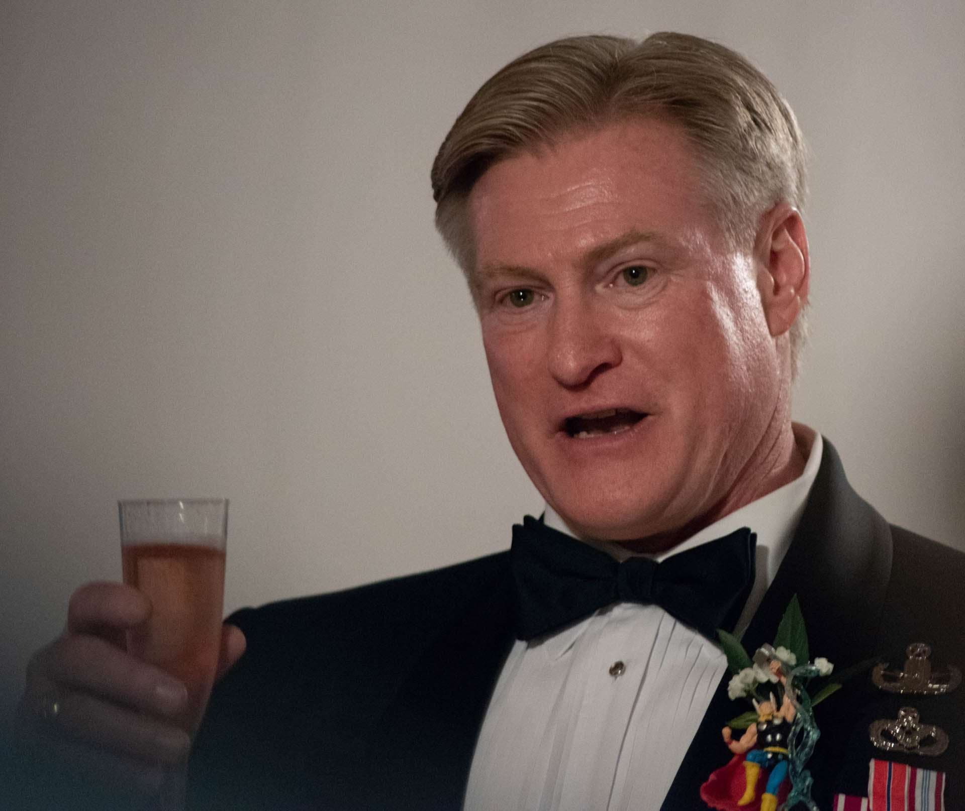 Man in a tuxedo and bow tie raising a drink in a toast, wearing a small superhero figurine boutonniere