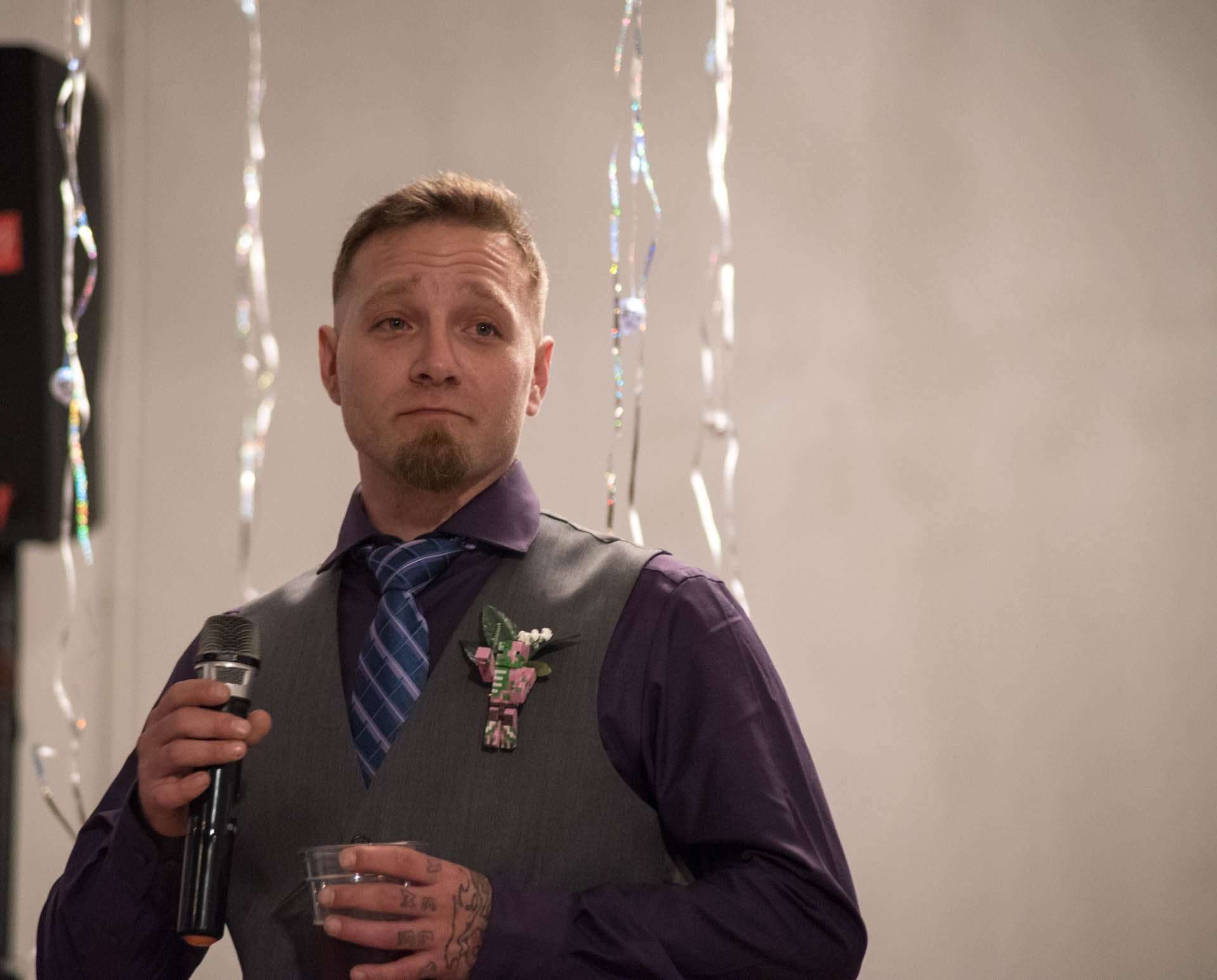 Man in purple shirt and gray vest with floral boutonniere holding a microphone and a cup, looking emotional during a toast