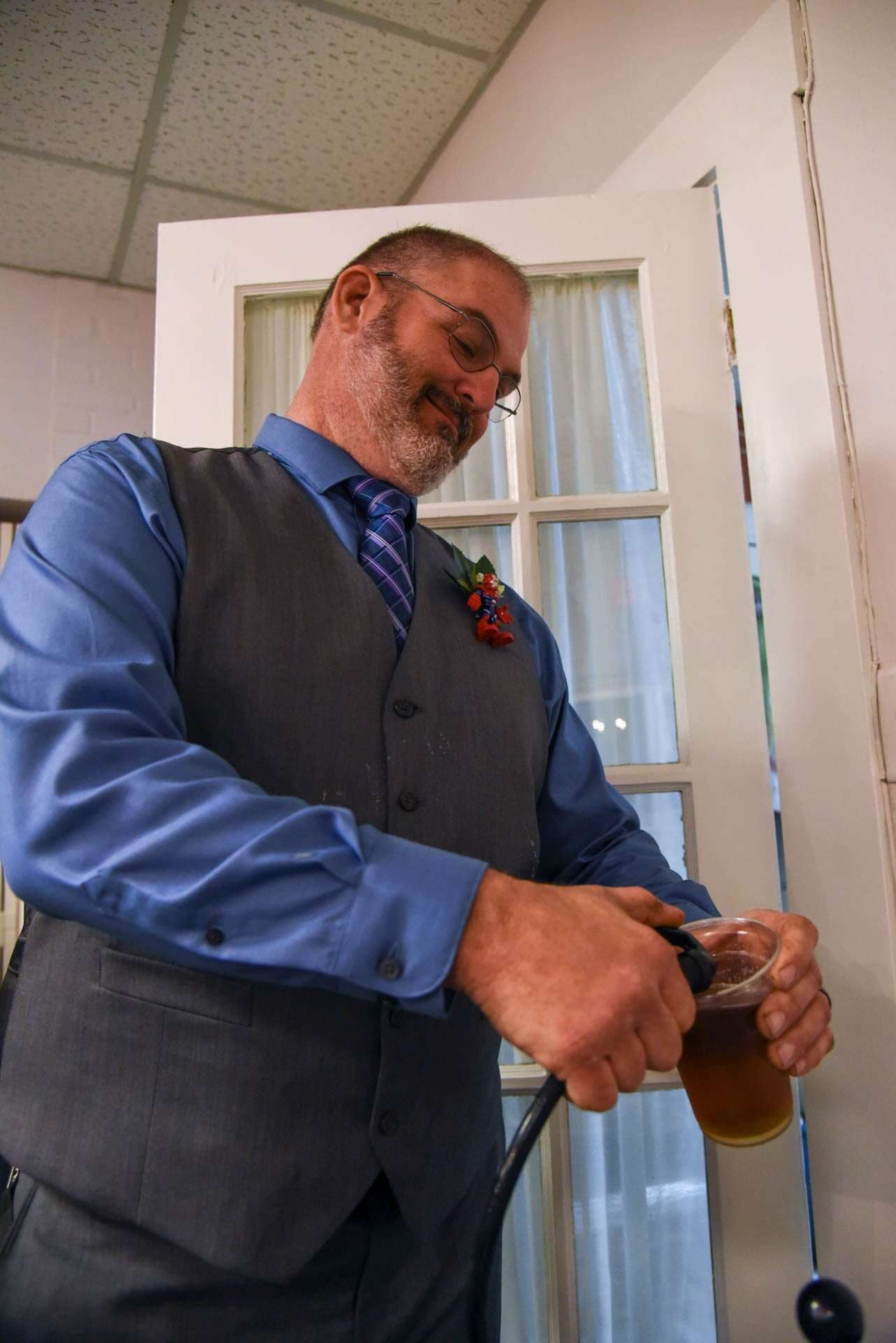 Bearded man in glasses, blue shirt, gray vest and floral boutonniere pours a beer from a tap into a plastic cup