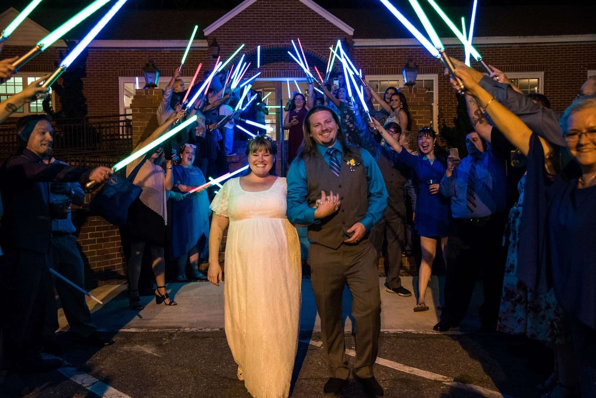 Couple walking through a tunnel of guests holding glowing lightsabers outside a brick building at night