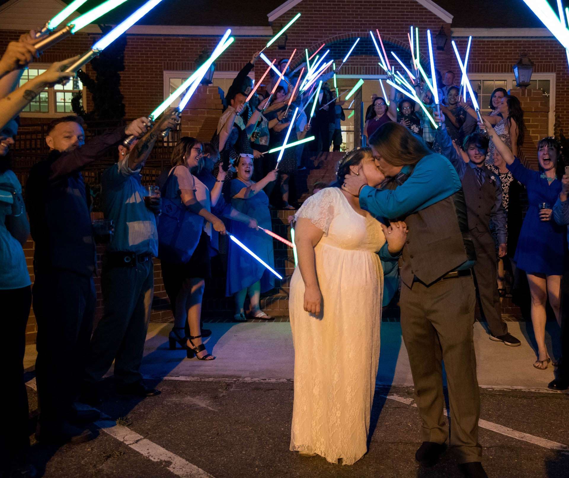 Couple kissing outside a brick building as guests form an archway of glowing lightsabers at night