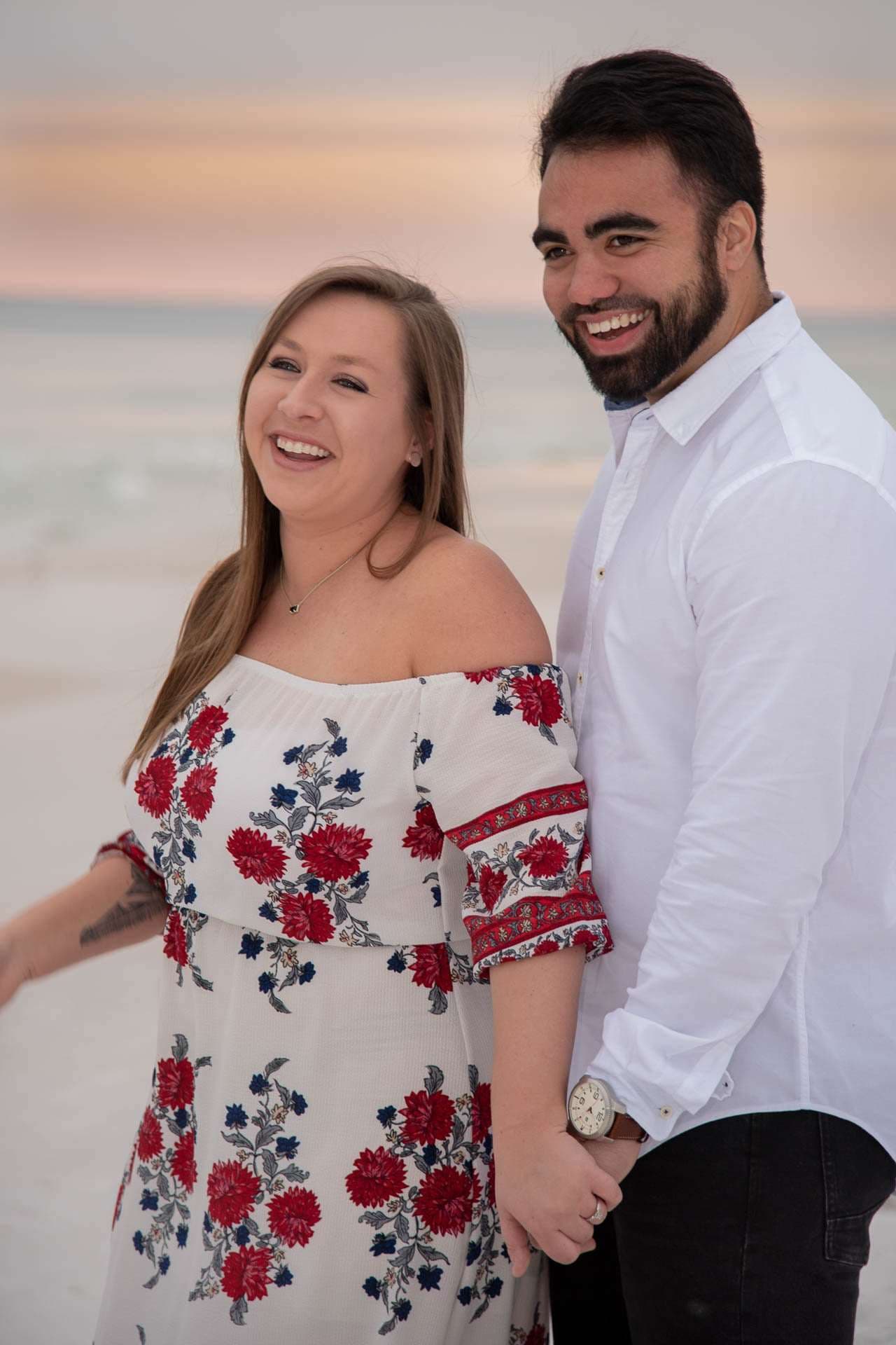Smiling couple holding hands on Grayton Beach at sunset, she in a red floral off-shoulder dress, he in a white shirt