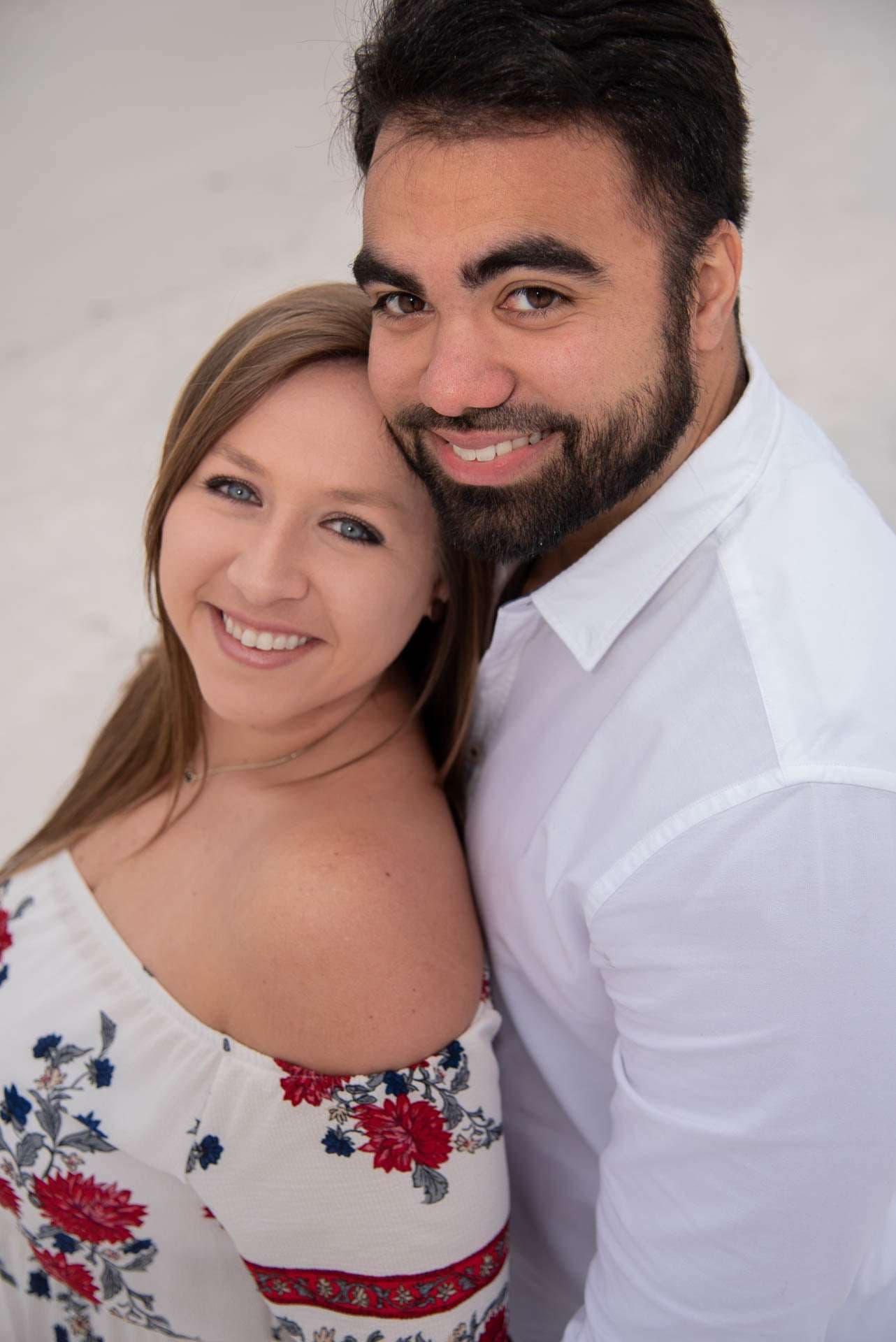 Smiling couple embracing cheek to cheek on white sand at Grayton Beach, she in a floral off-shoulder top, he in a white shirt