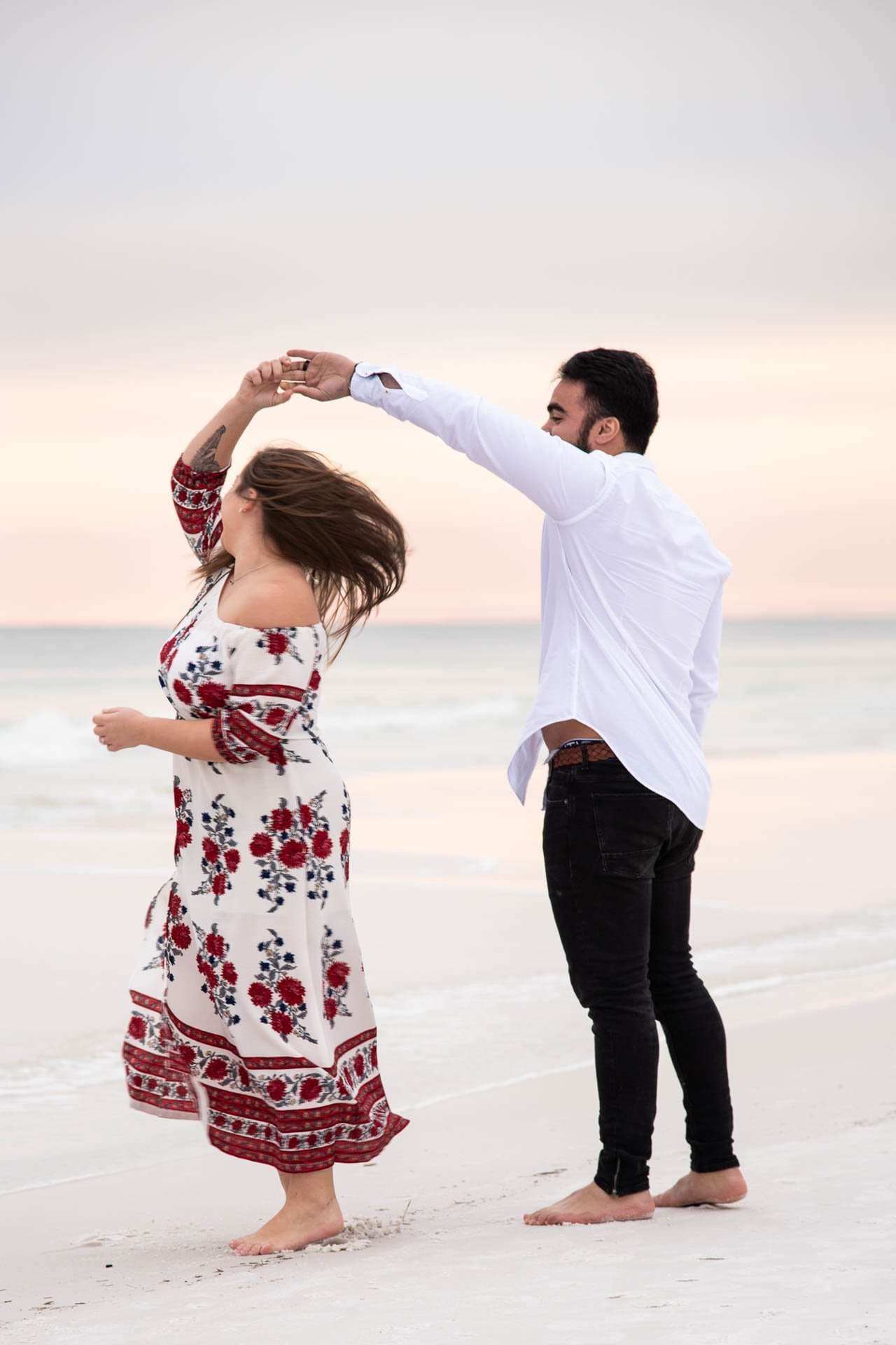Couple dancing barefoot on Grayton Beach at sunset, man twirling a woman in a red floral dress