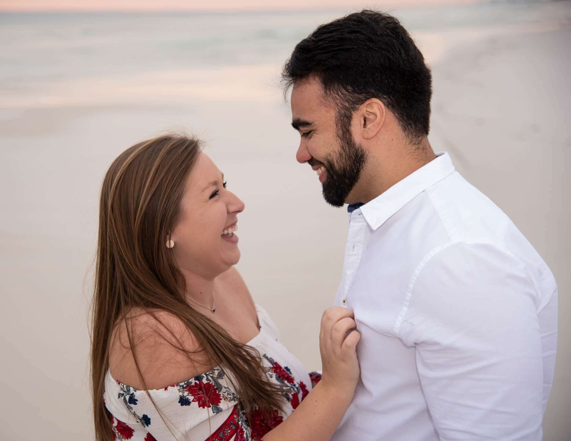 Couple laughing face to face on the sand at Grayton Beach, she tugs his white shirt, soft pastel dusk behind them
