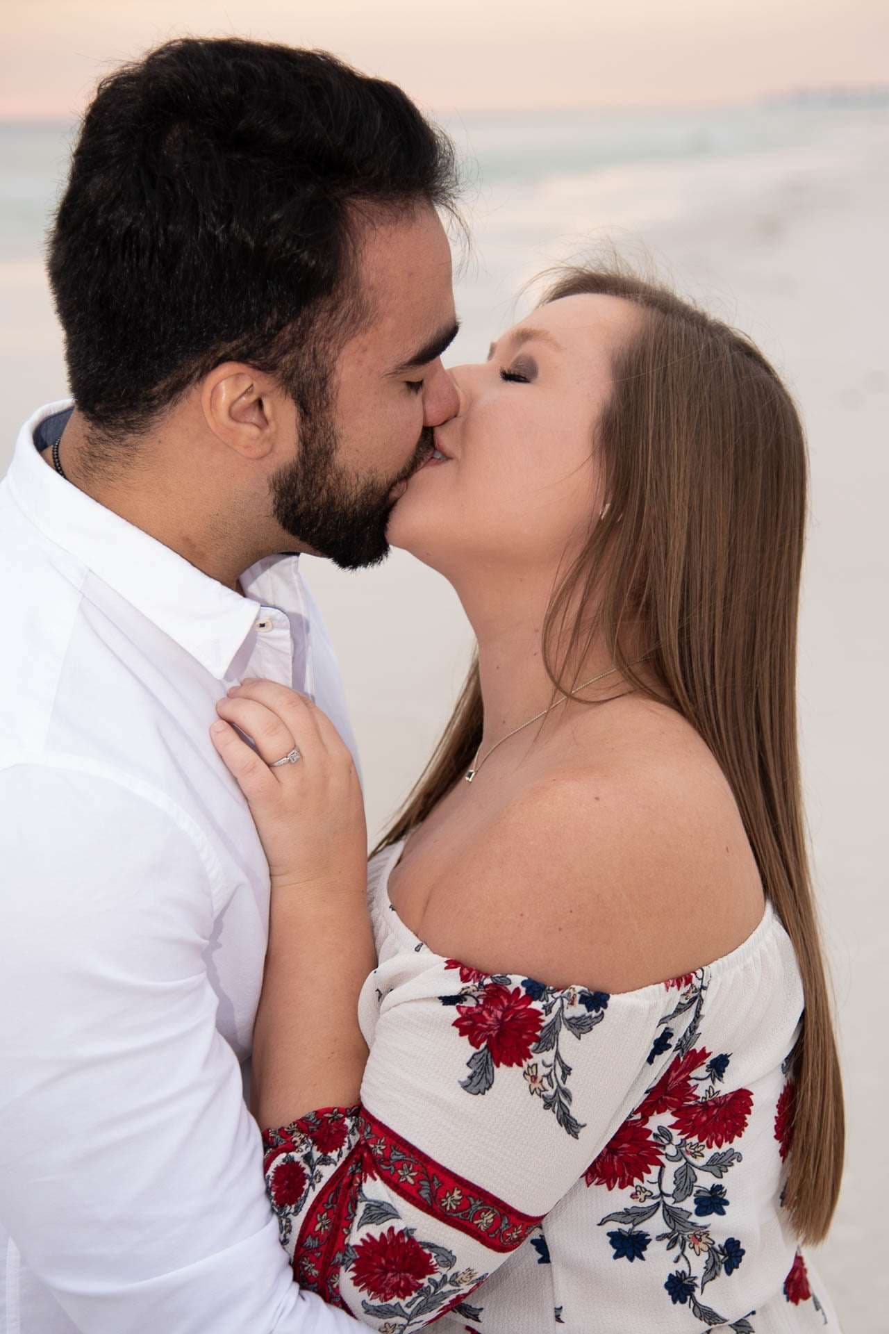 Couple kissing on the beach at sunset, her hand with an engagement ring resting on his white shirt
