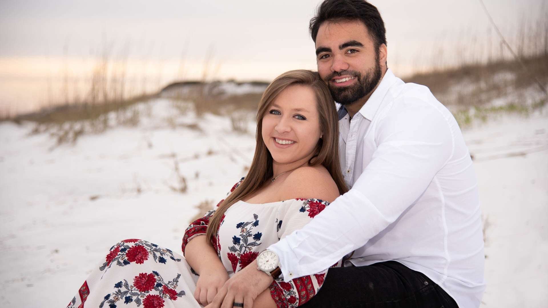 Smiling couple embracing on white sand dunes at Grayton Beach, she wears a red floral dress, he a white shirt