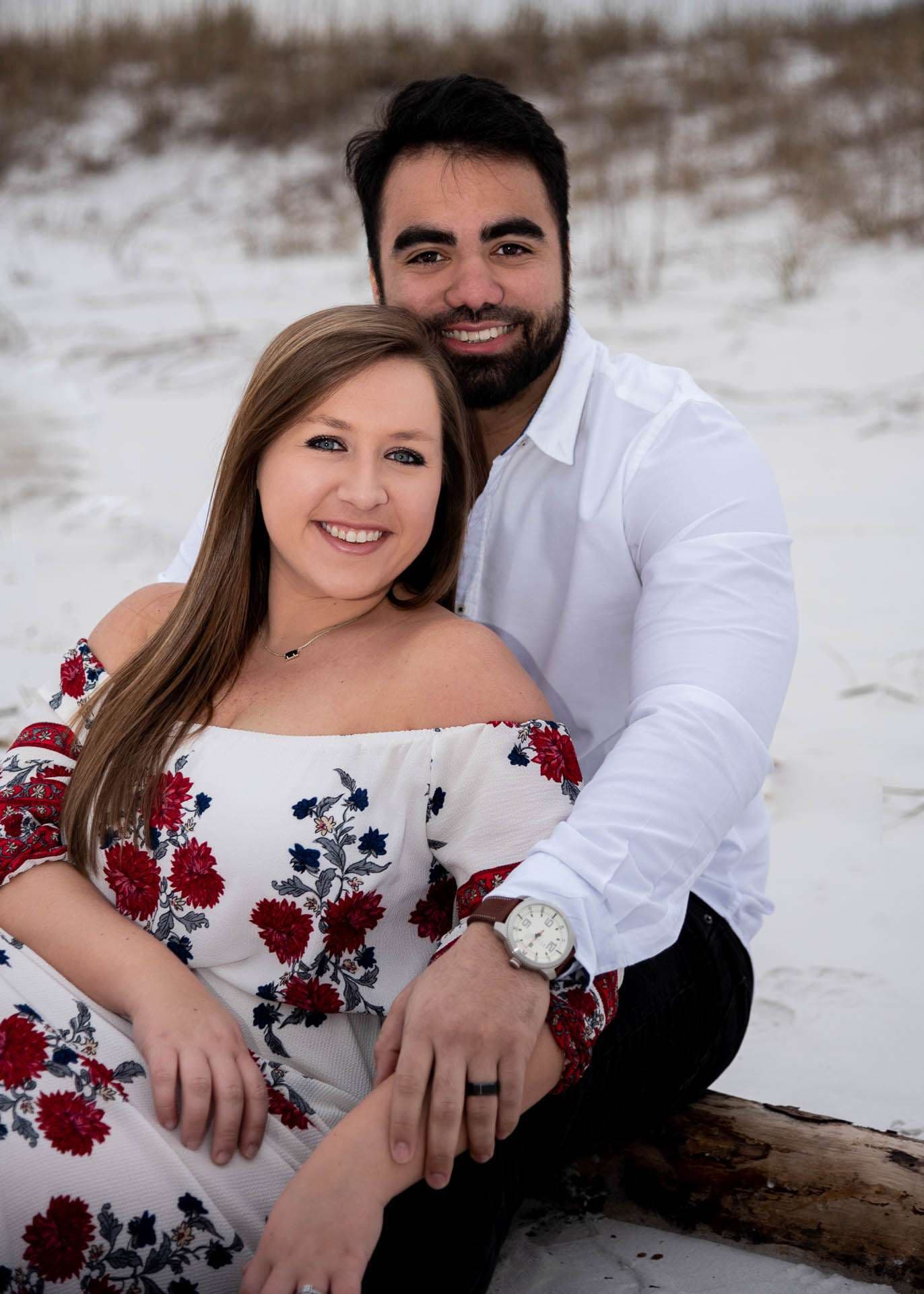 Smiling couple embracing on a log at Grayton Beach, the woman in a red floral off-shoulder dress, dune grass behind
