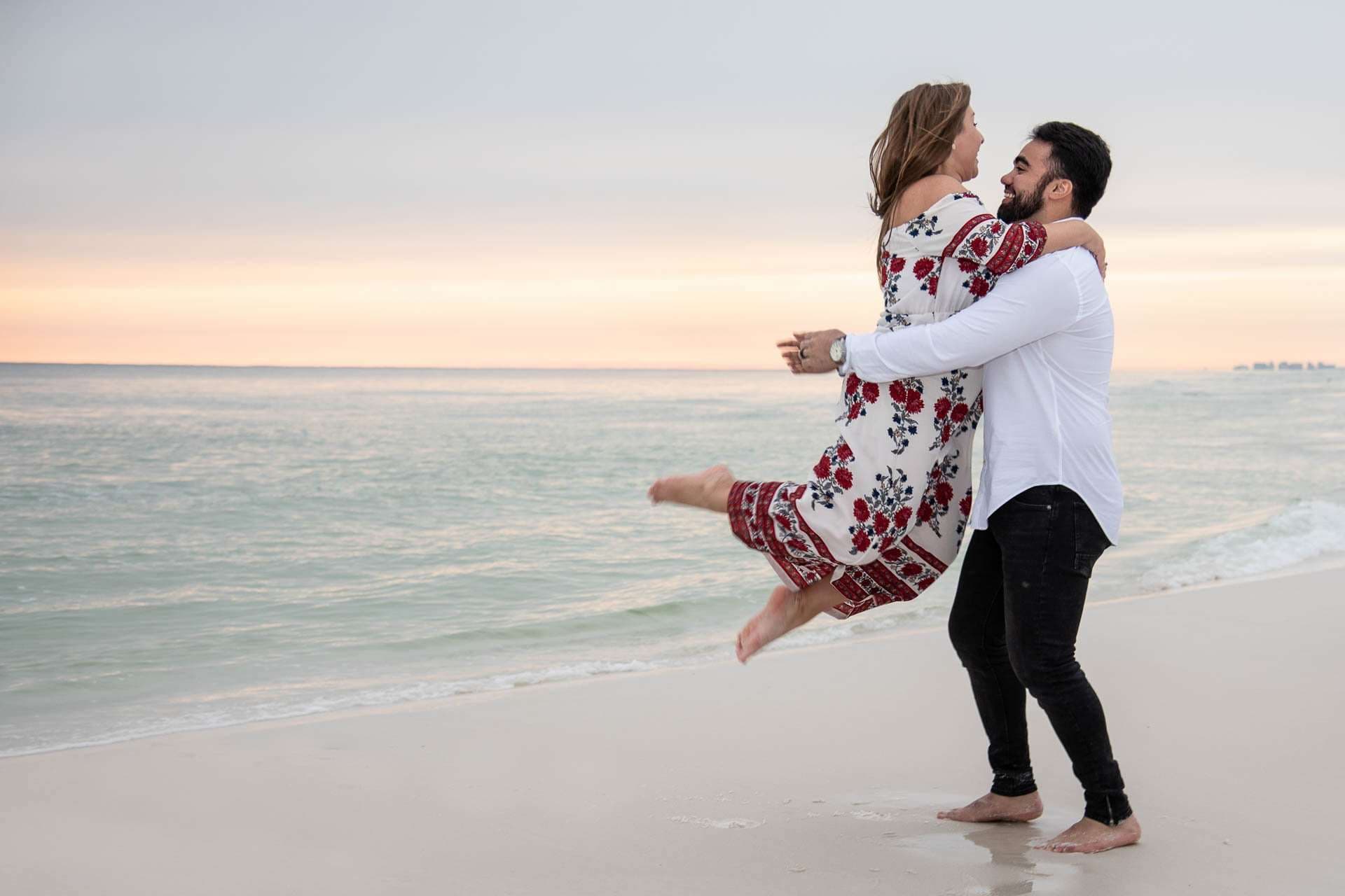 Man in white shirt lifting and spinning a laughing woman in a floral dress on Grayton Beach at sunset