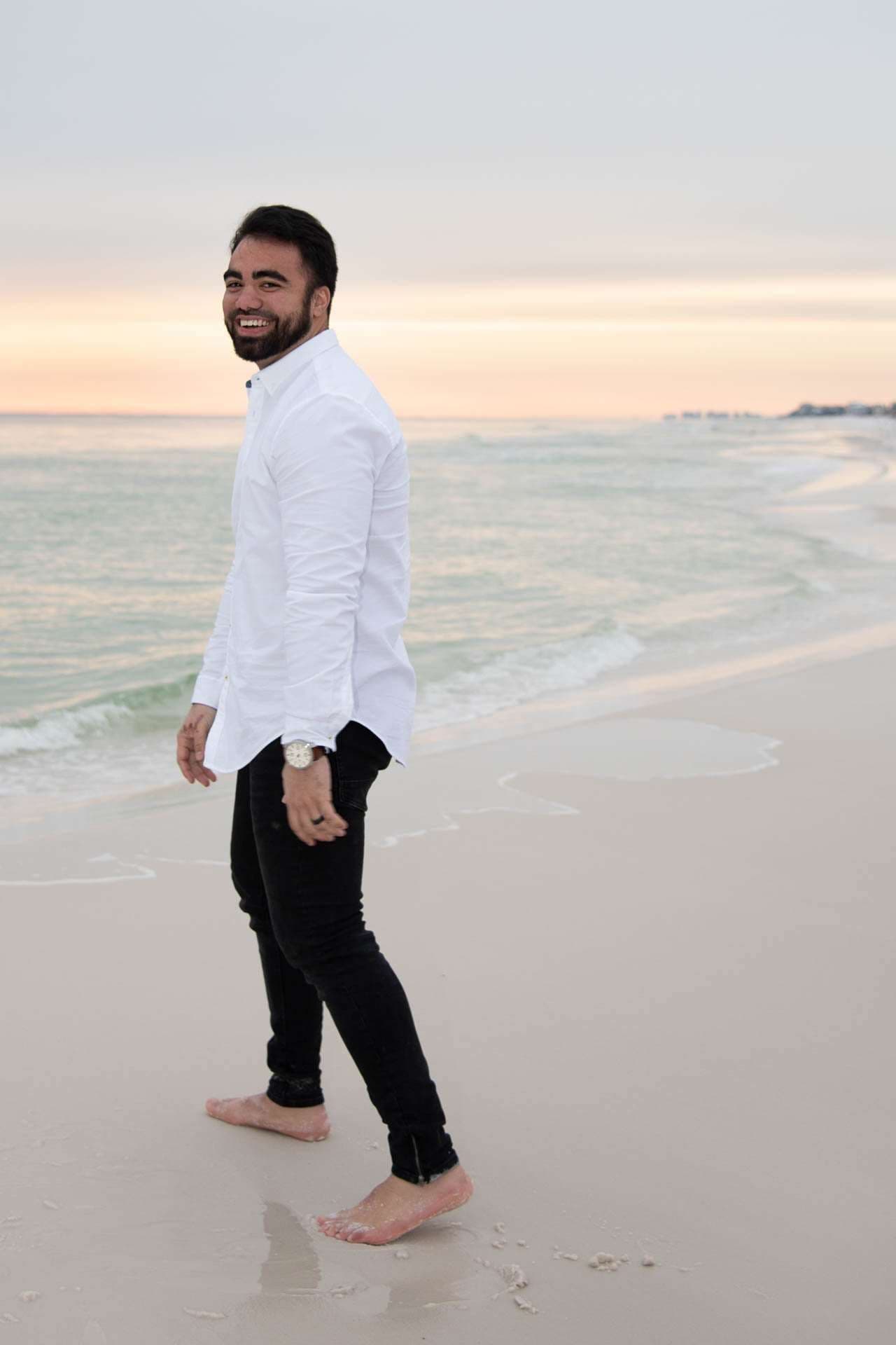 Smiling bearded man in white shirt and black pants walking barefoot along the shoreline at Grayton Beach at sunset