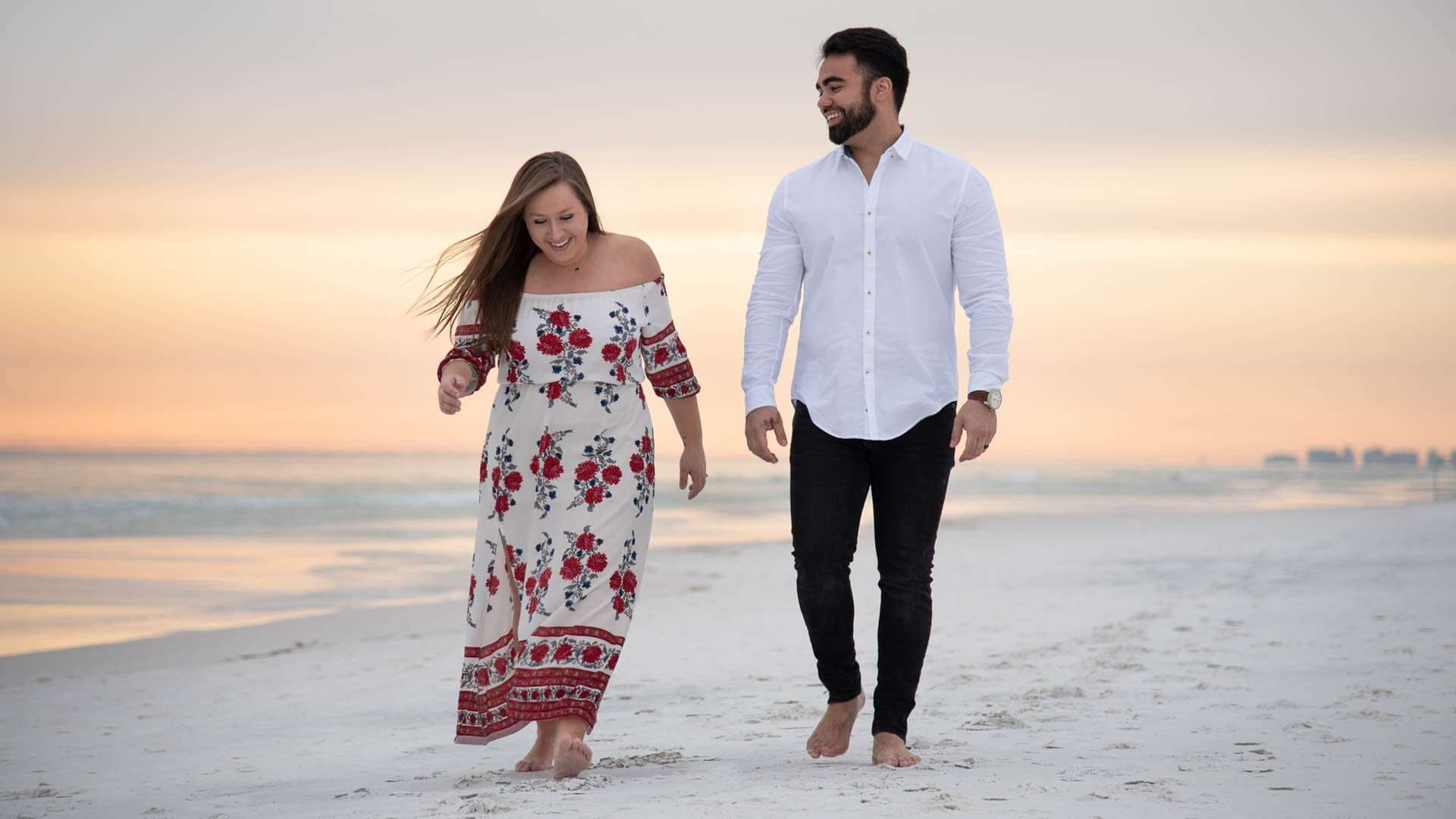 Couple walking barefoot and laughing on Grayton Beach at sunset, she in a red floral dress, he in a white shirt