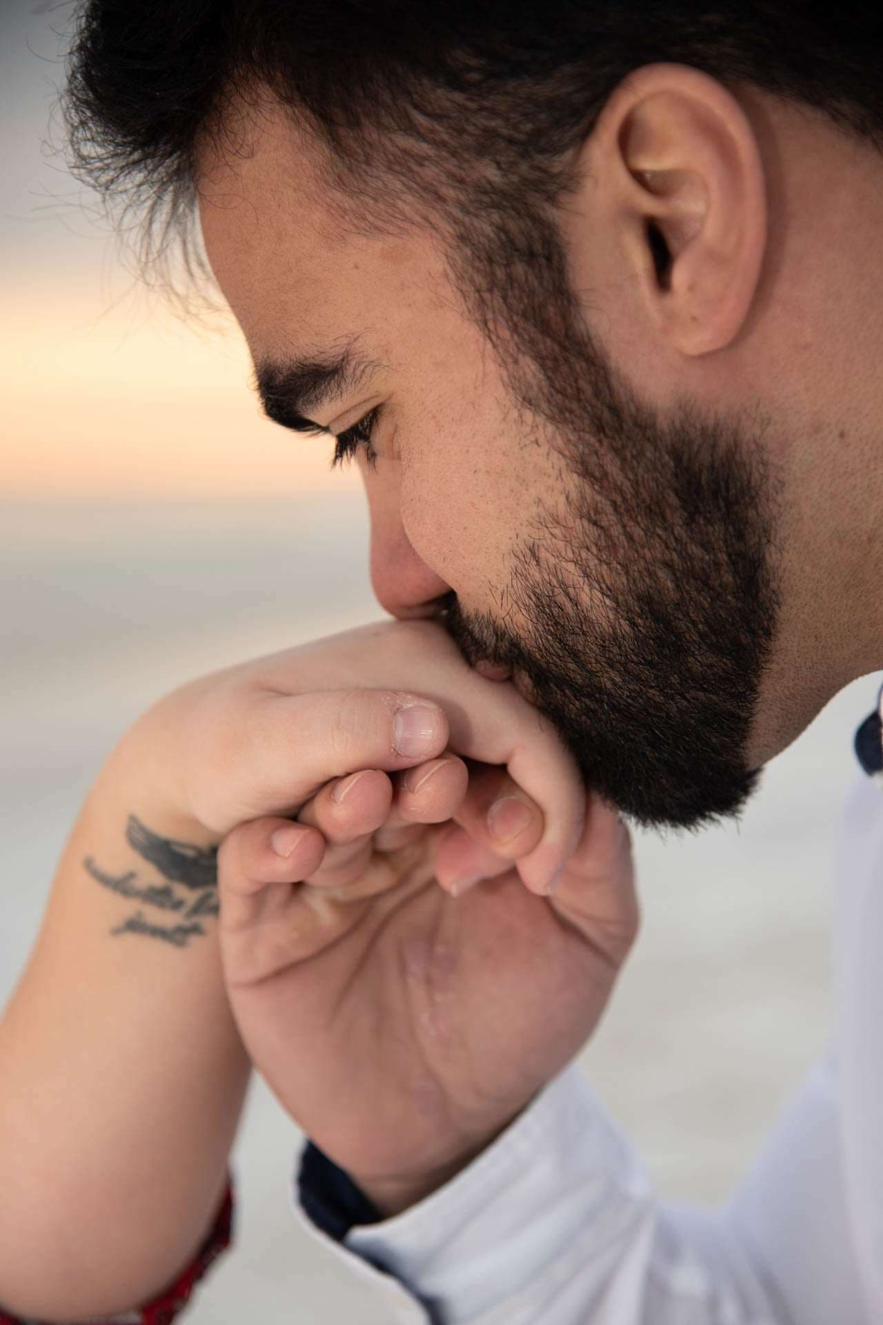 Bearded man in a white shirt kisses a hand with a small wrist tattoo, soft sunset glow behind him