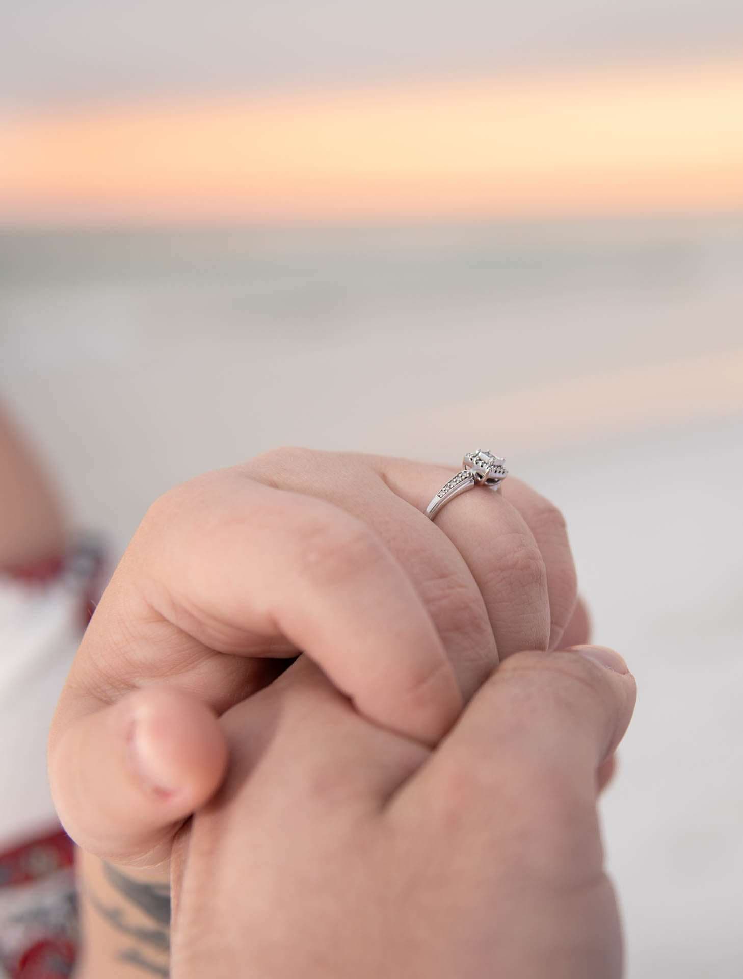 Close-up of clasped hands with a diamond engagement ring on the beach at Grayton Beach during a pastel sunset