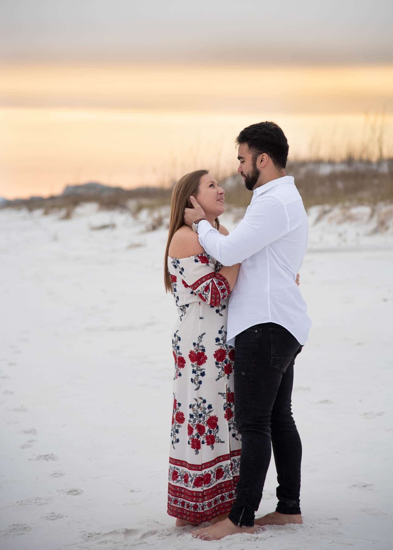 Couple embracing barefoot on white sand at Grayton Beach at sunset, she in a red floral maxi dress, he in a white shirt