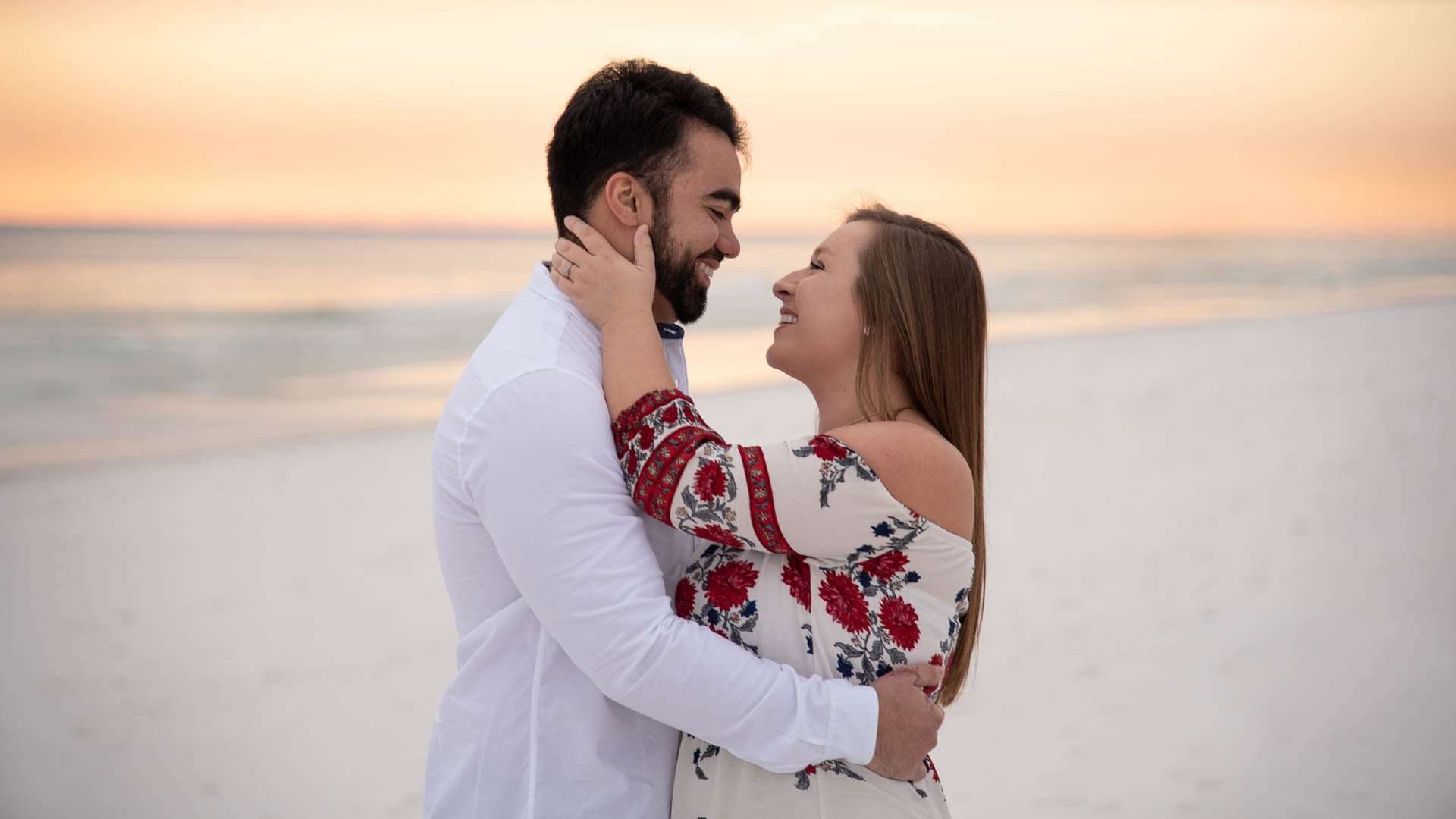 Couple embracing and smiling on Grayton Beach at sunset, woman in a red floral embroidered top, man in white shirt