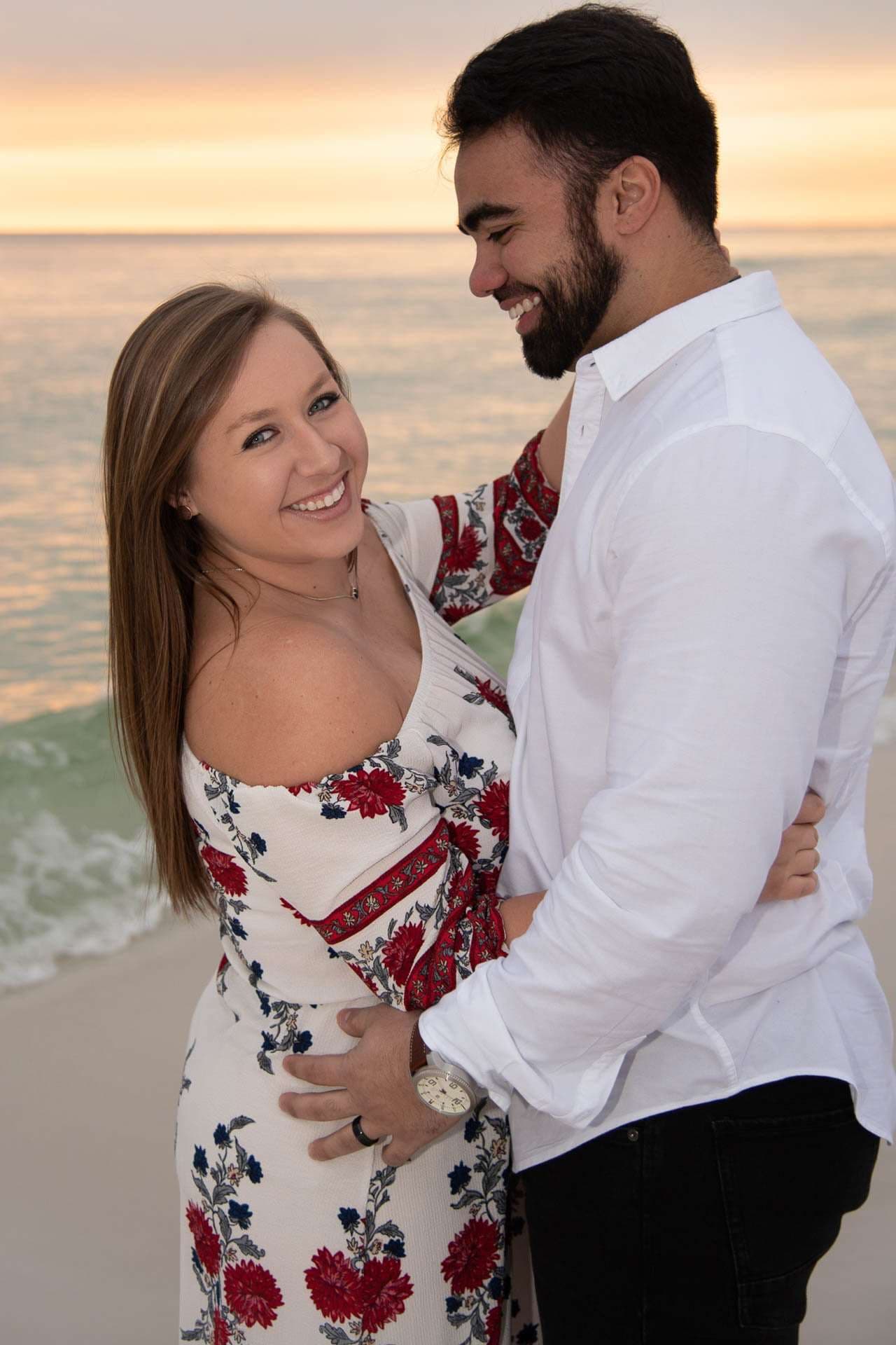 Smiling couple embracing on Grayton Beach at sunset, she wears a red floral dress and he a white shirt