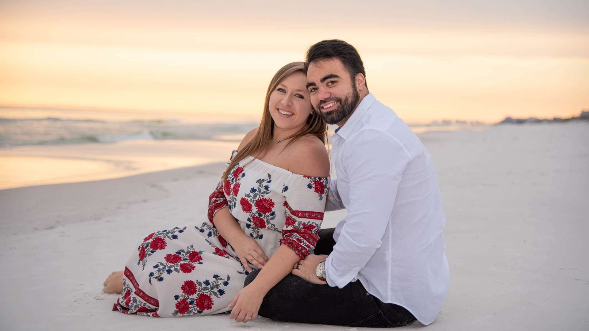 Smiling couple sitting close together on white sand at sunset, woman in a red floral off-shoulder dress, man in white shirt
