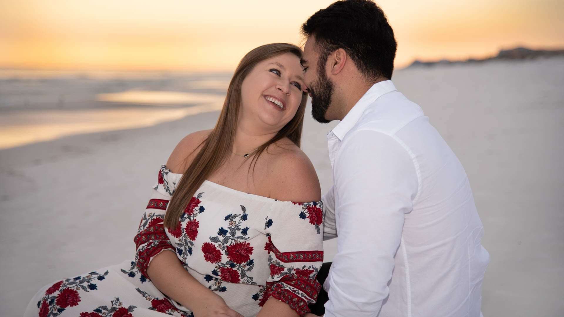 Couple sharing a tender moment on Grayton Beach at sunset, she laughs in a red floral off-shoulder dress, he wears white