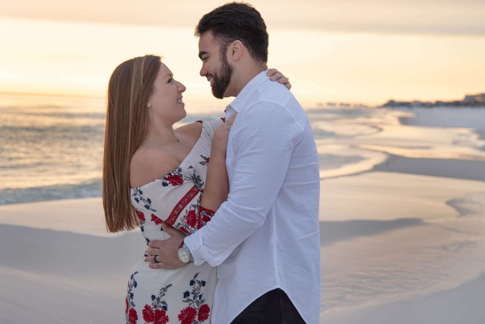 Couple embracing and smiling at each other on Grayton Beach at sunset, woman in a red floral dress