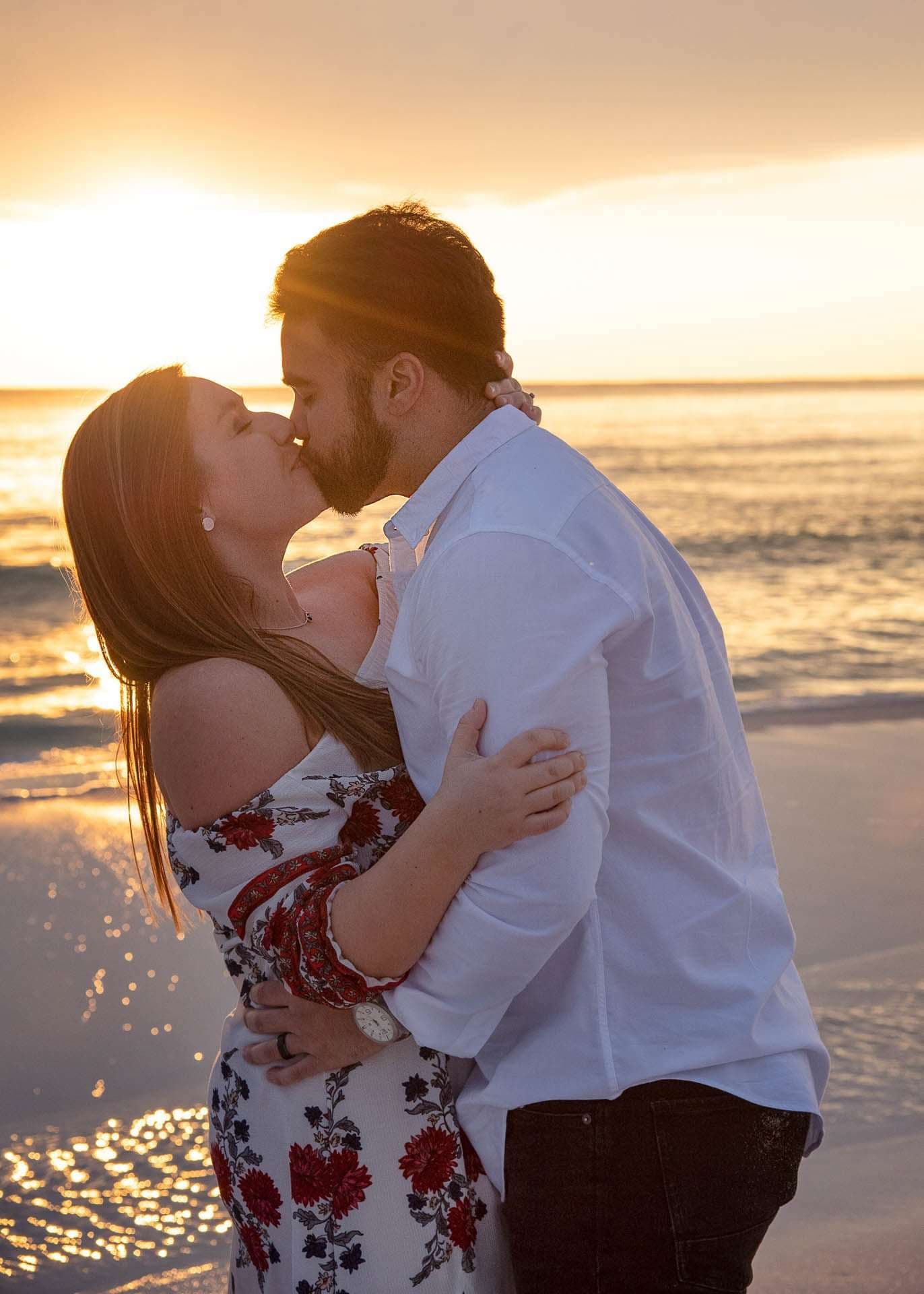 Couple kissing and embracing on Grayton Beach at sunset, woman in a red floral dress, man in a white shirt