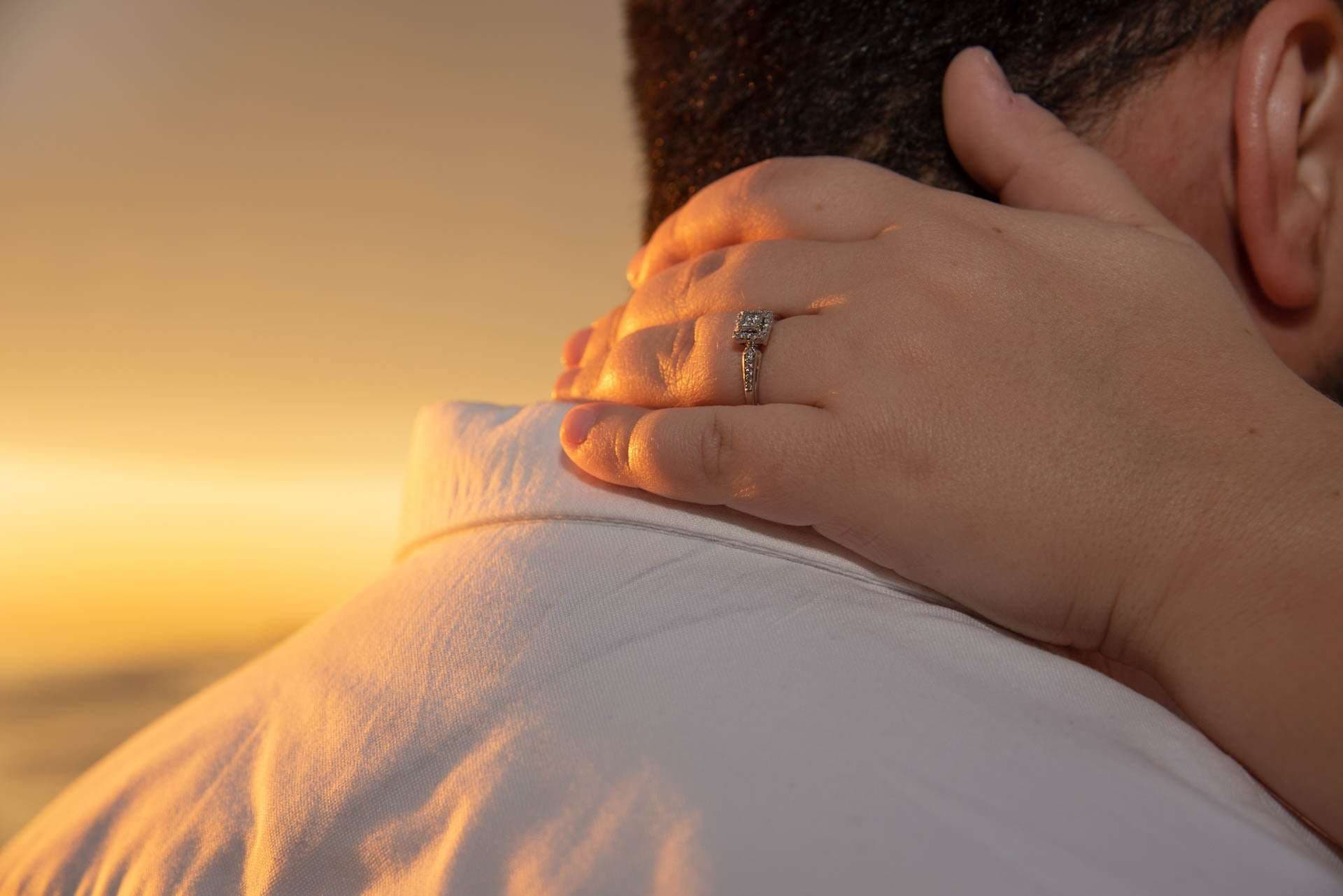 Close-up of a hand with a diamond engagement ring resting on a partner's neck at golden sunset, Grayton Beach