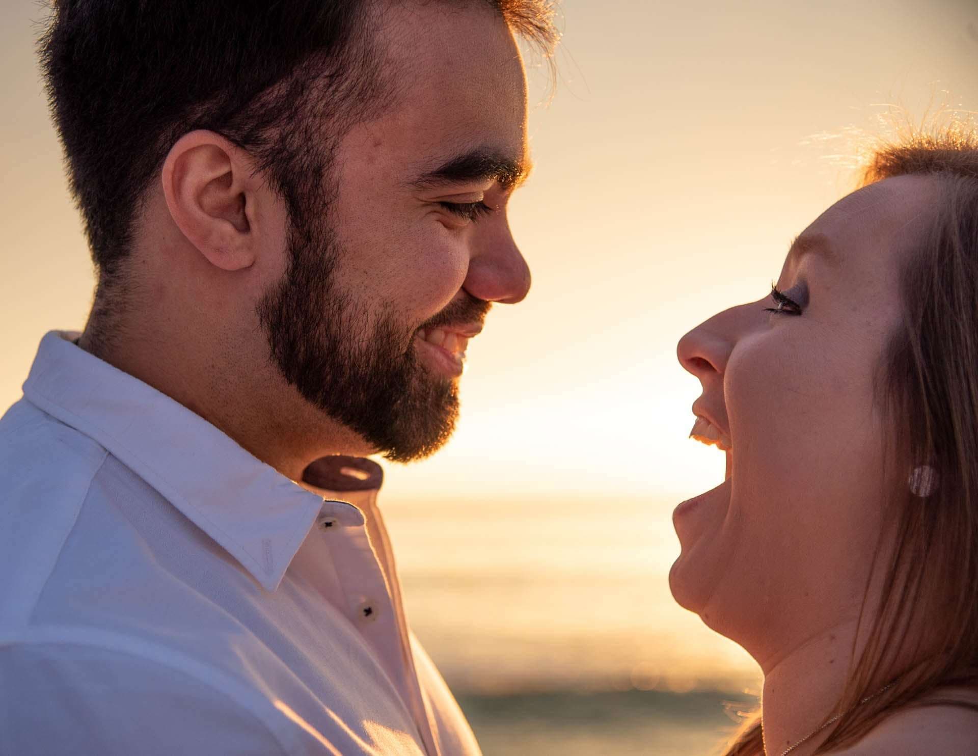 Smiling bearded man in a white shirt faces a laughing woman in profile against a golden sunset at Grayton Beach