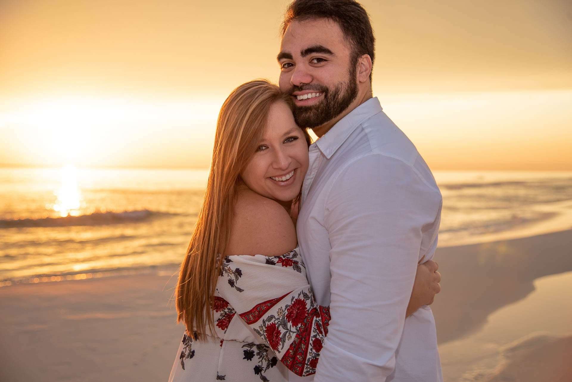 Smiling couple embracing on Grayton Beach at sunset, woman in a floral off-shoulder top, man in a white shirt