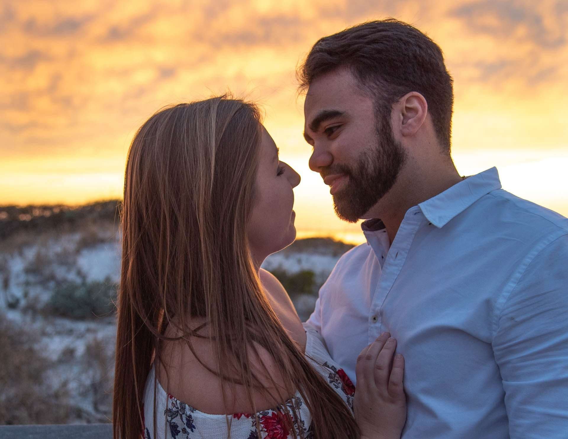 Couple almost touching noses at sunset on Grayton Beach, with dunes and a glowing orange sky behind them