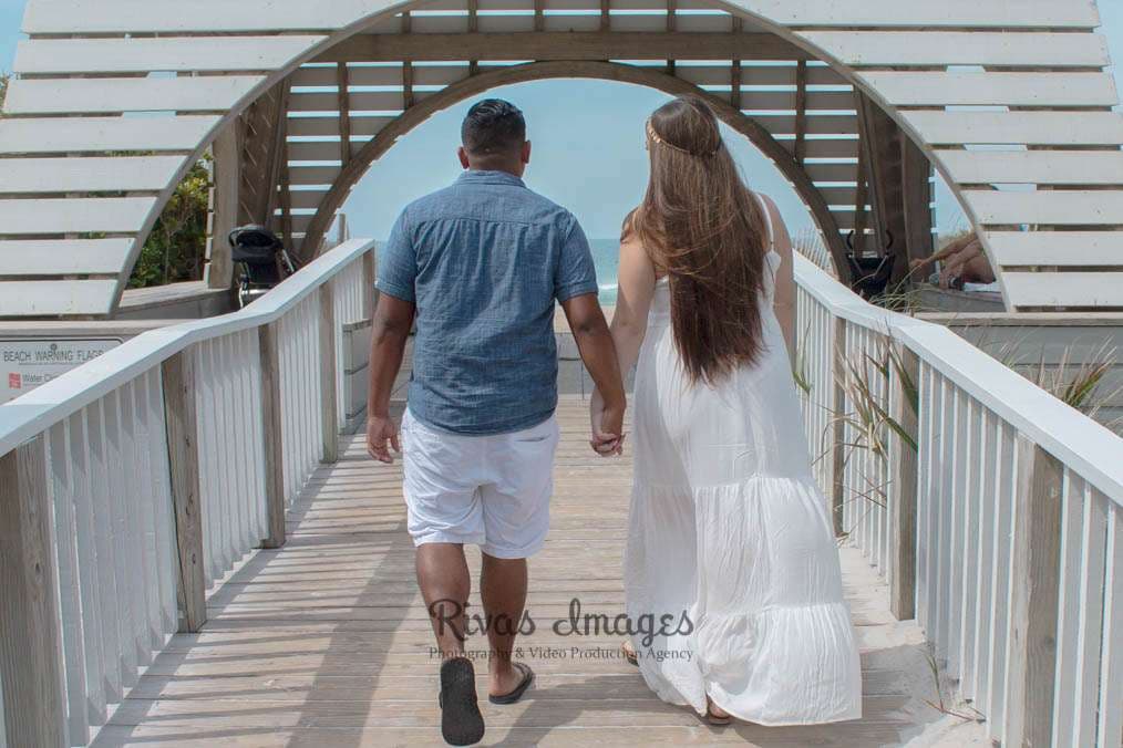 Couple holding hands and walking away on a white wooden boardwalk toward an arched gateway and the ocean in Seaside, Florida